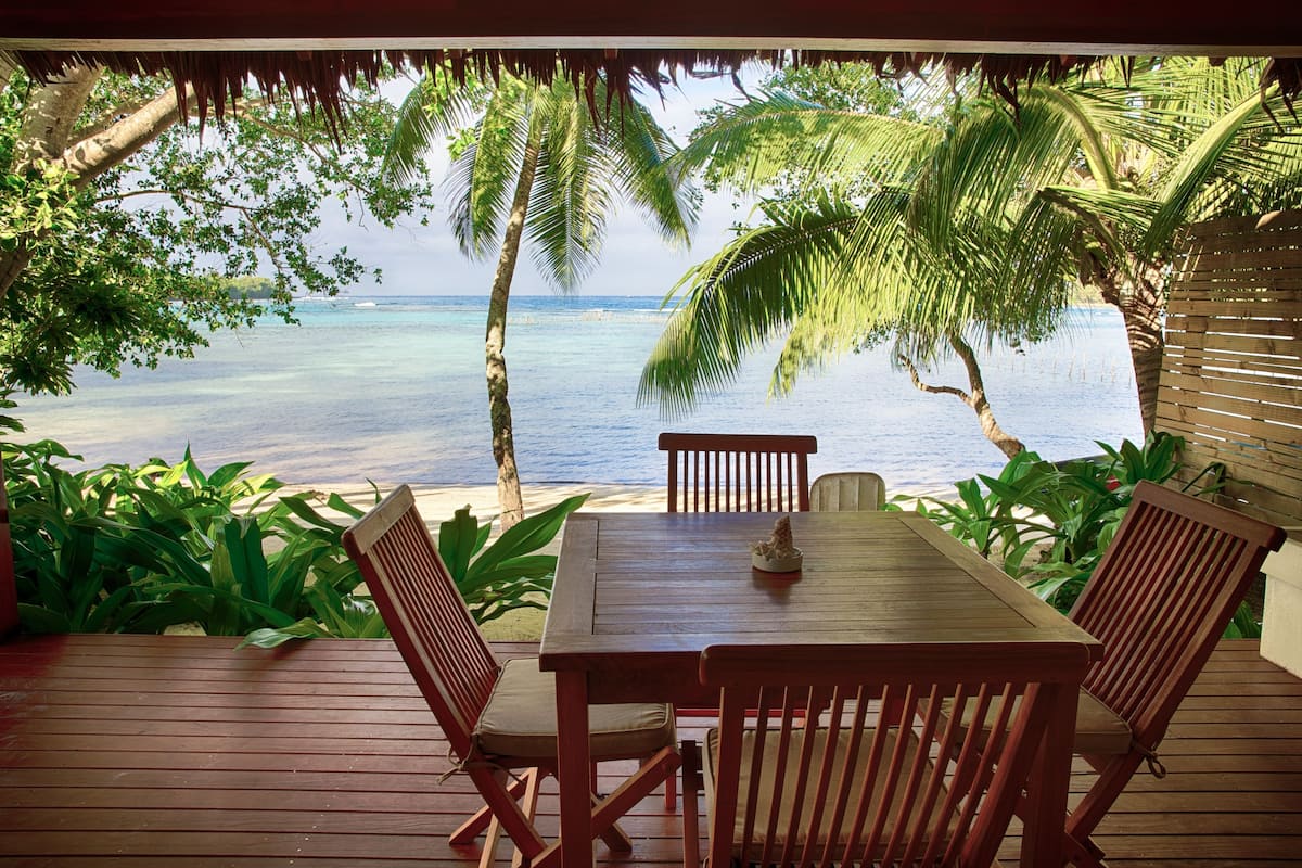 A wooden deck with a table and chairs overlooking a beach with palm trees and clear blue water.