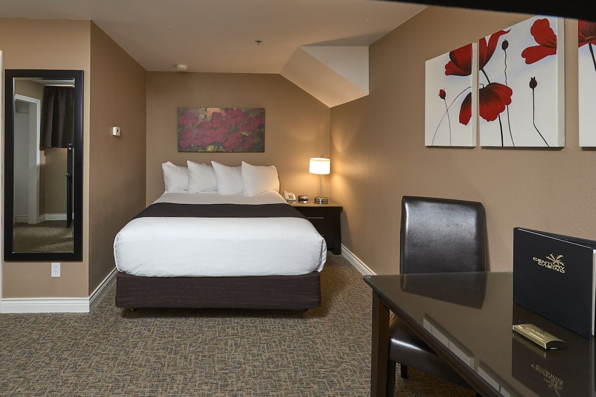 A hotel room with a bed, a desk, a chair, a lamp, and artwork on the walls.