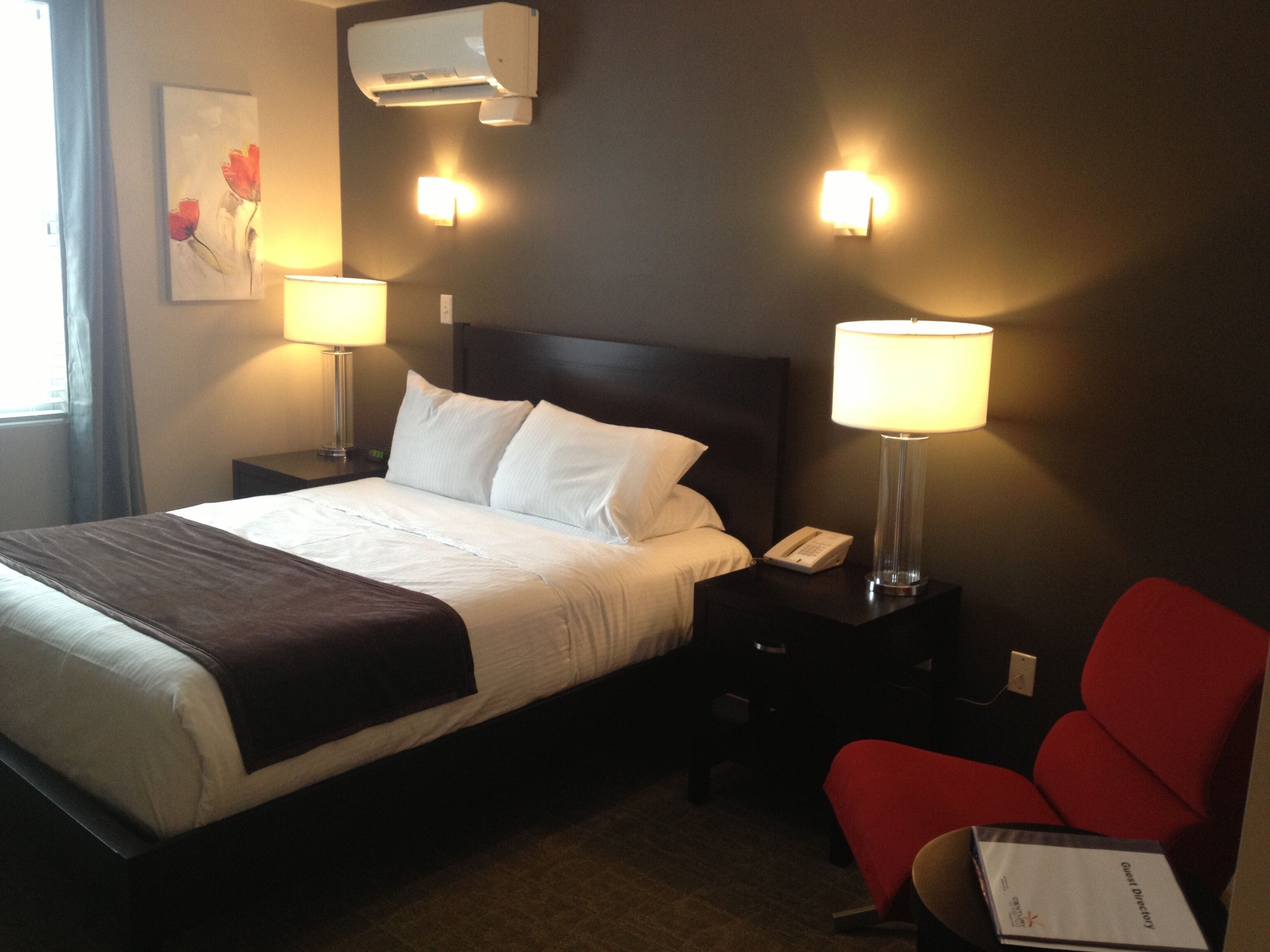 Habitación de hotel con cama, dos lámparas de noche, mesita de noche con teléfono, una silla roja y aire acondicionado instalado en la pared.