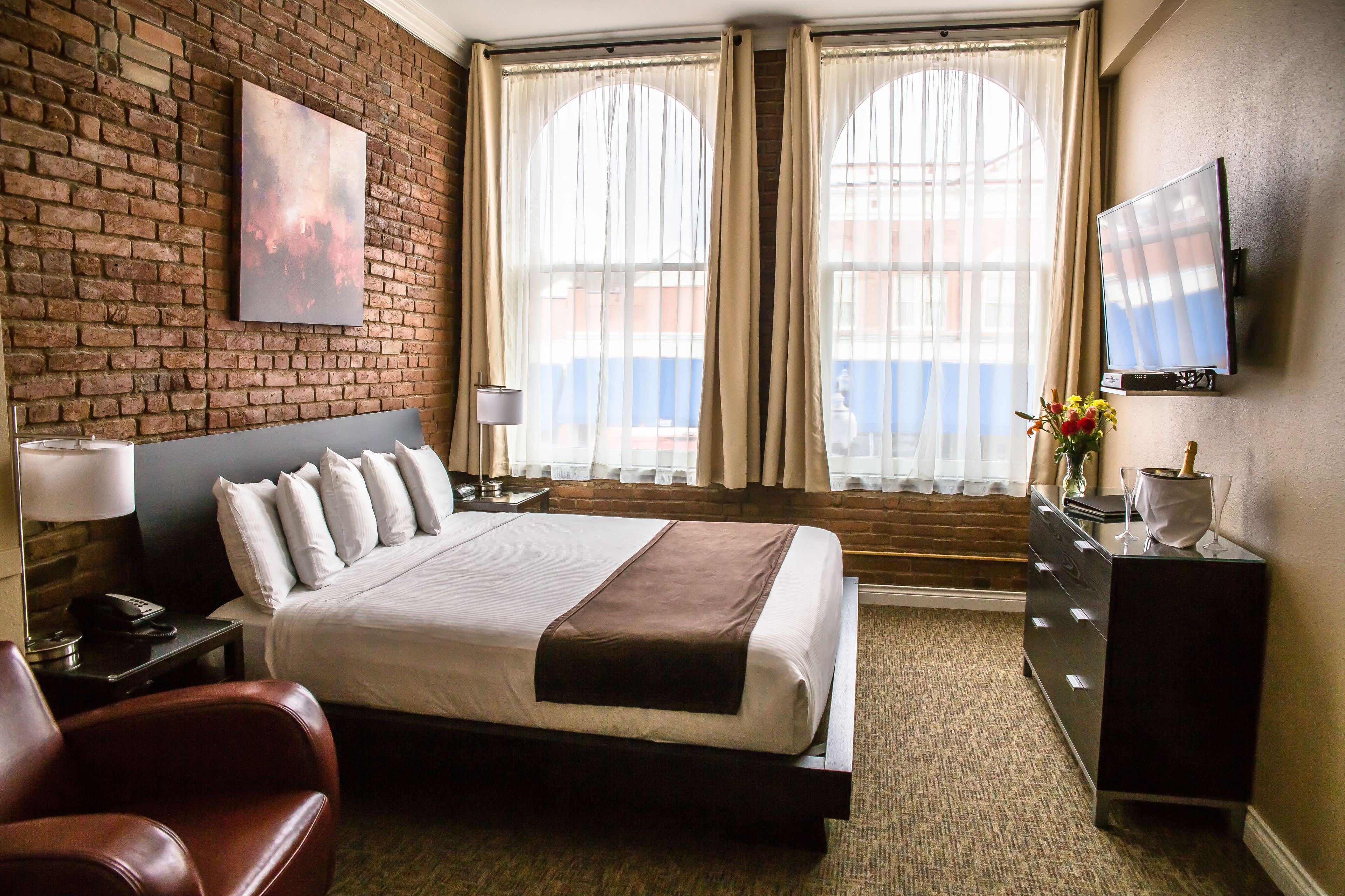 Una habitación de hotel con una cama grande, televisión, escritorio con lámpara y pared de ladrillo.