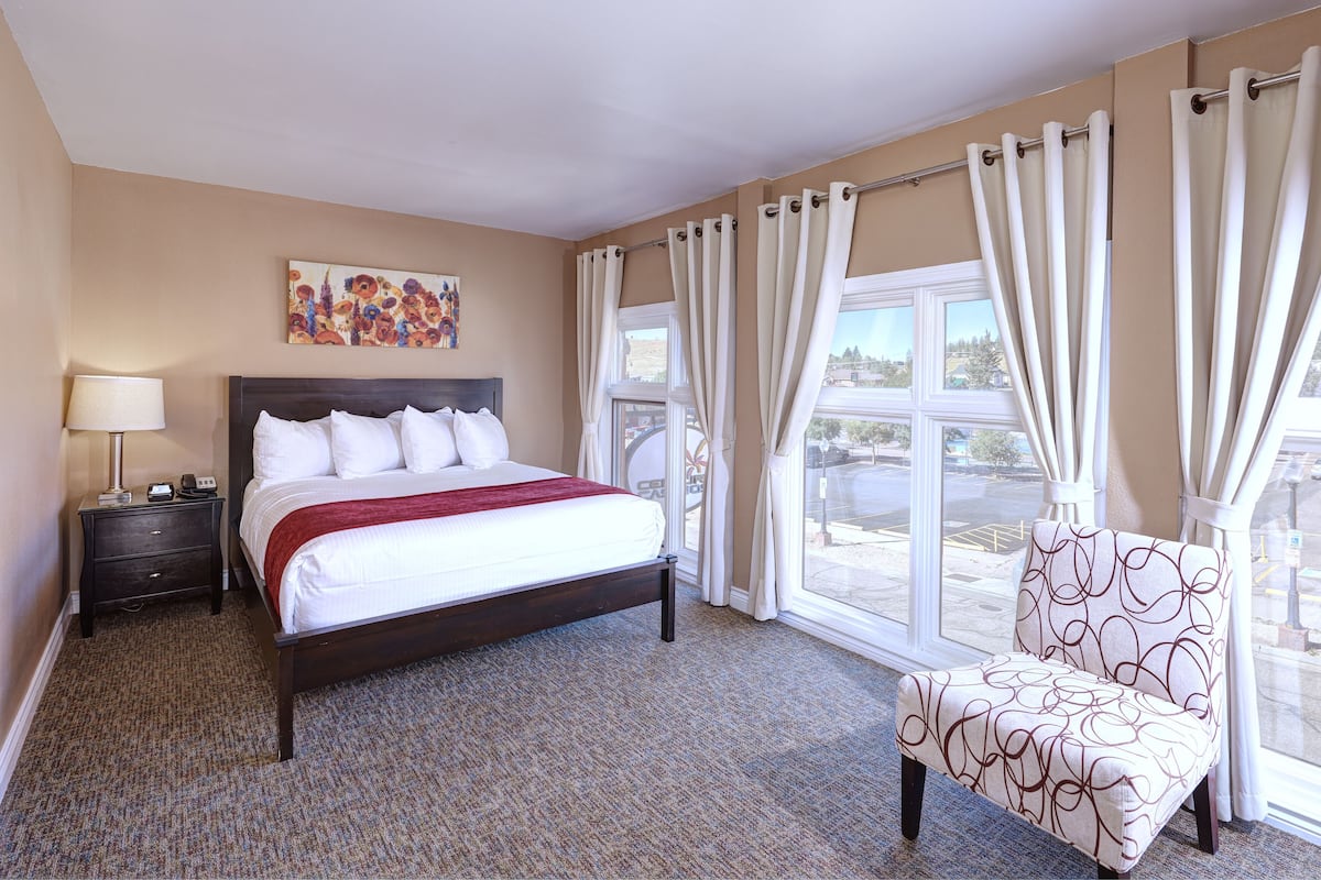 A hotel room with a bed, a chair, a nightstand, a lamp, a painting, and large windows with curtains.