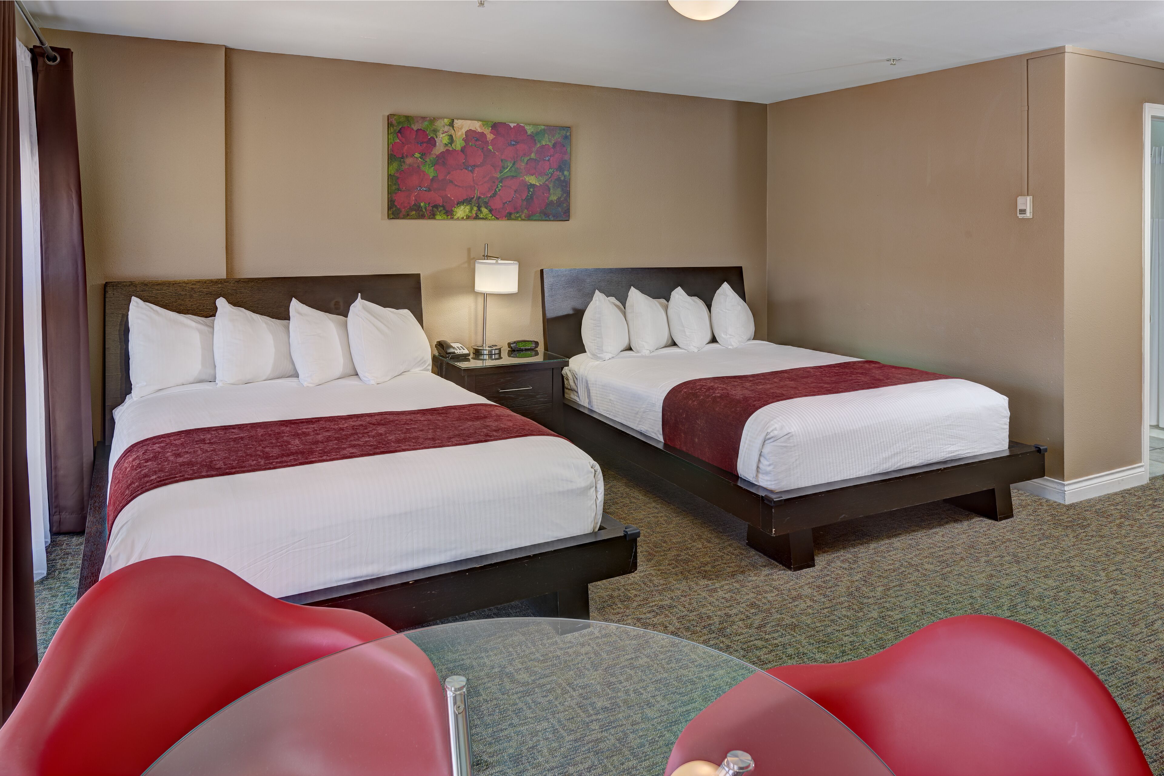 A hotel room with two beds, a red chair, a small table, and a painting on the wall.