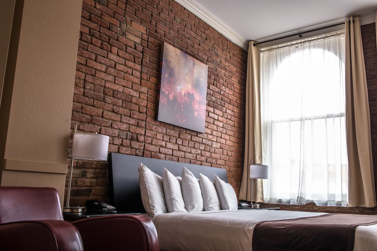 A hotel room with a brick wall, a large window with curtains, a bed with multiple pillows, and a desk with a lamp.