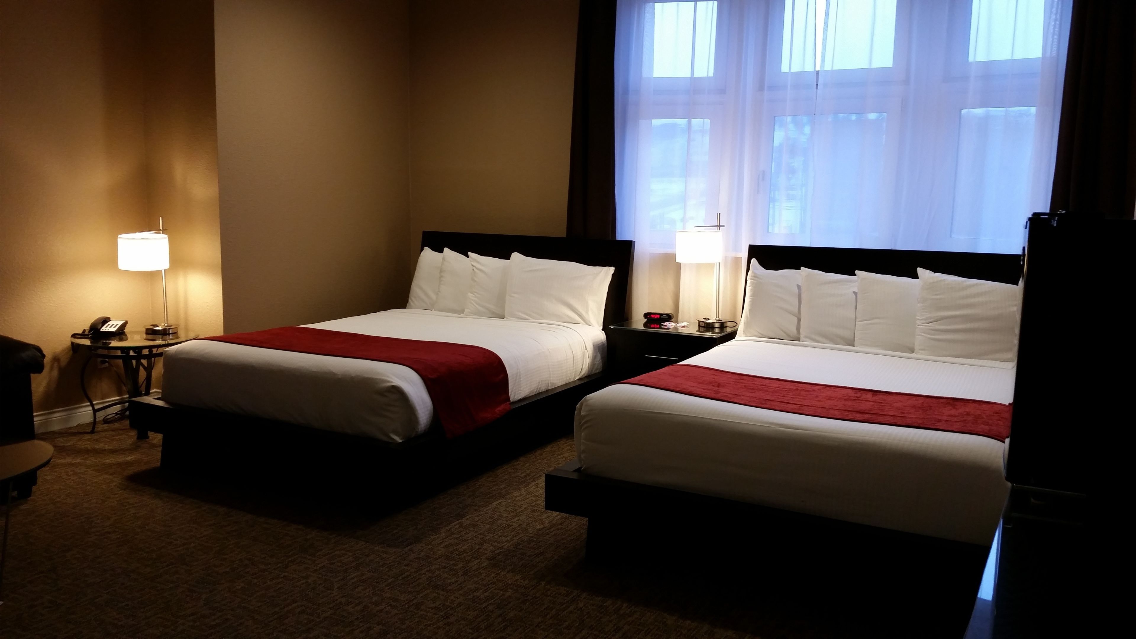 Une chambre d’hôtel avec deux lits, chacun agrémenté de draps blancs et de touches de couleur rouge. On y trouve des tables de chevet avec des lampes et un téléphone. La chambre est dotée d’un sol recouvert de moquette et de grandes fenêtres avec des rideaux légers.