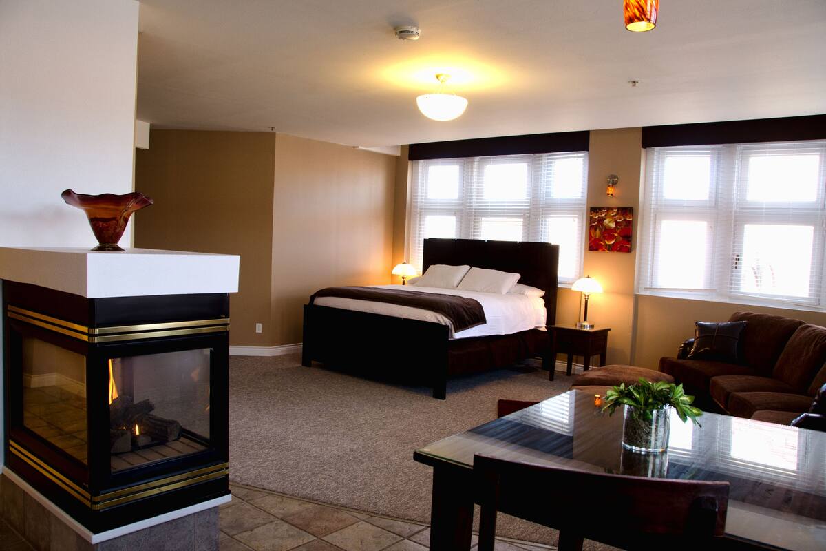 A bedroom with a bed, bedside lamps, a fireplace, a sofa, and a dining table.