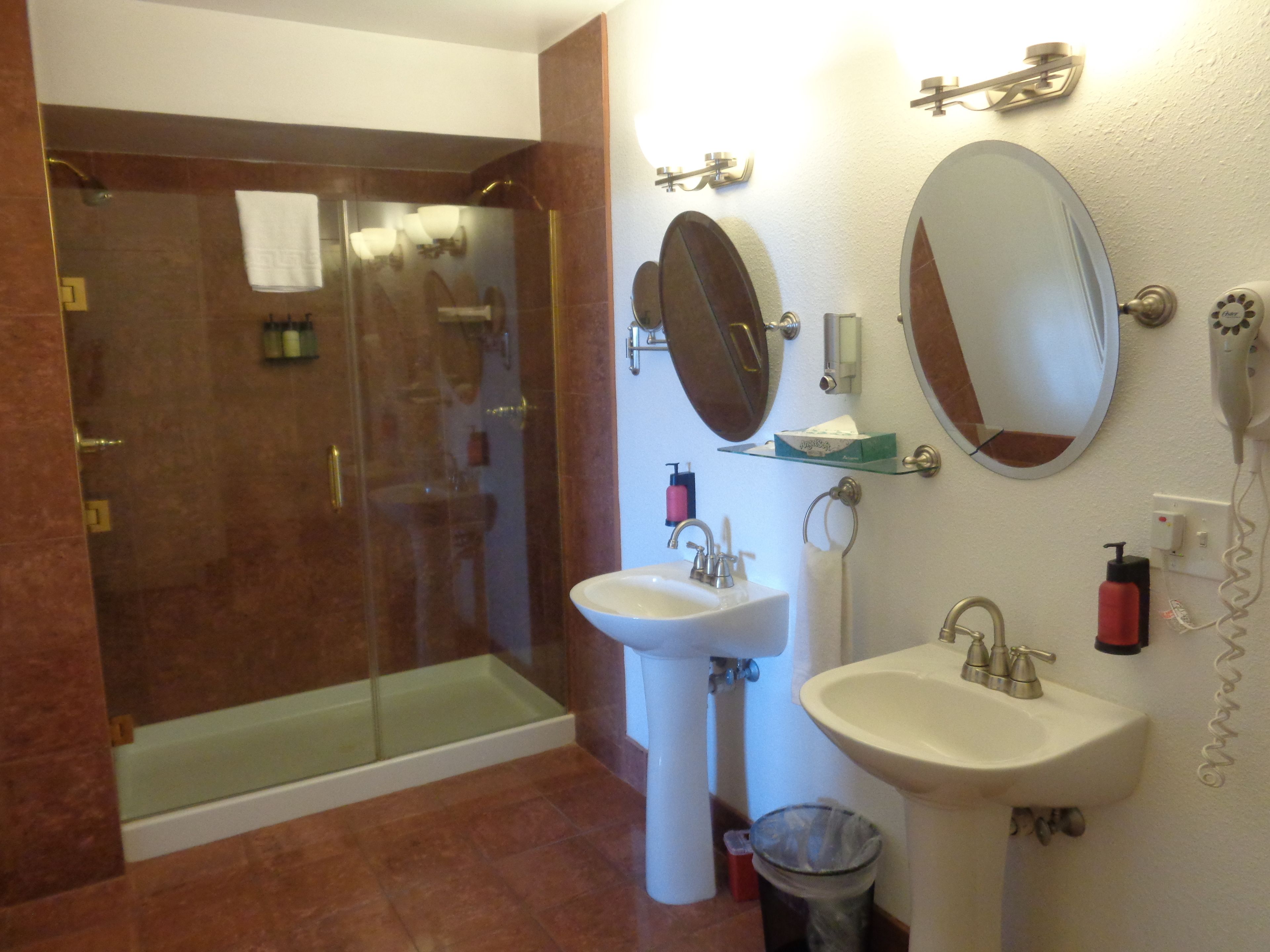 A bathroom with a glass-enclosed shower, two sinks, a mirror, and toiletries on the wall.