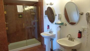 A bathroom with a glass-enclosed shower, two sinks, a mirror, and toiletries on the wall.