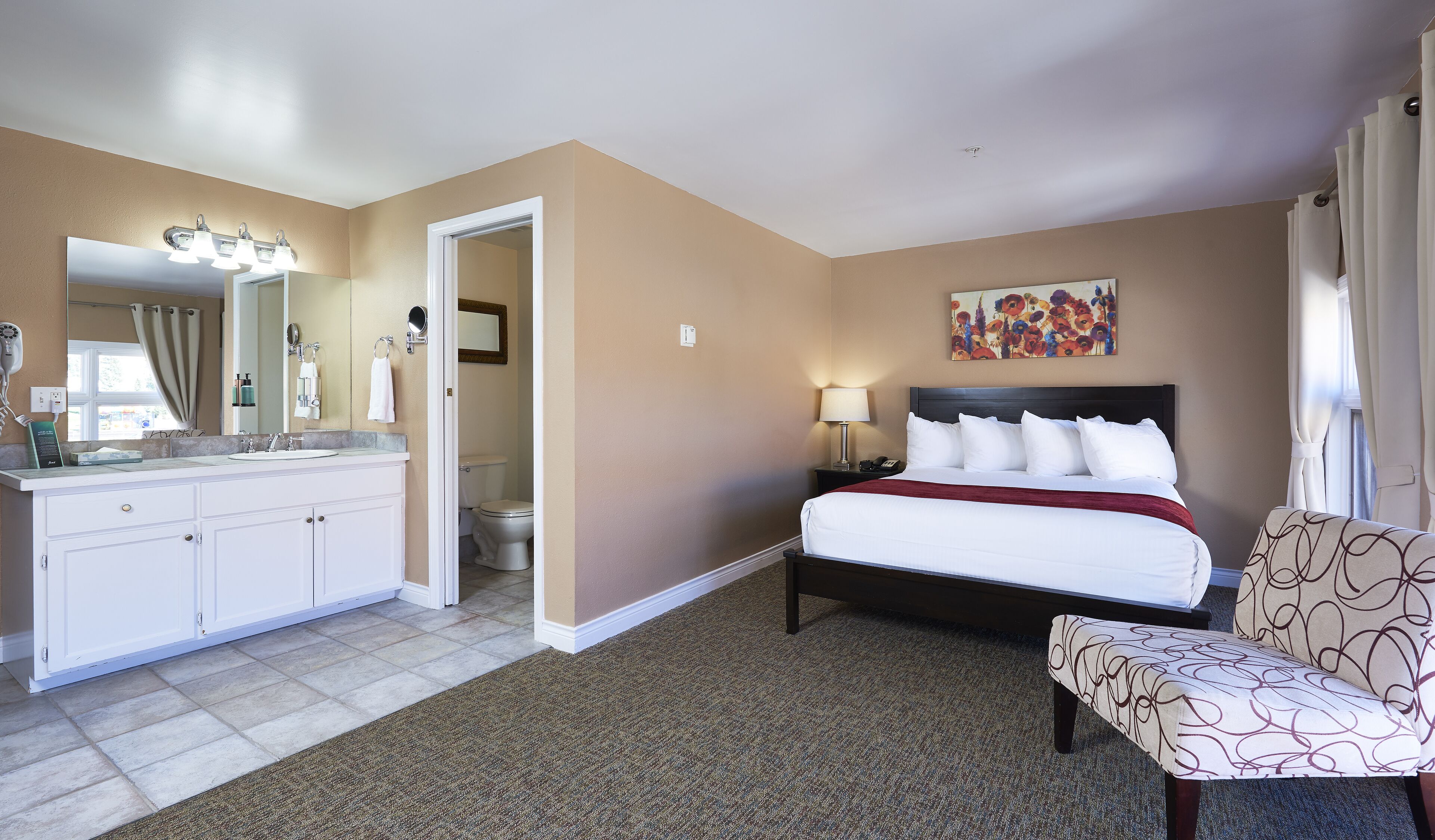 A hotel room with a large bed, a sitting area, a bathroom with a vanity, and a painting on the wall.