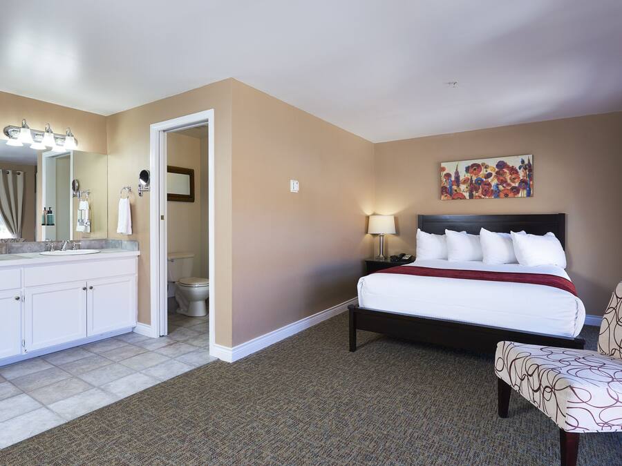 A hotel room with a large bed, a sitting area, a bathroom with a vanity, and a painting on the wall.