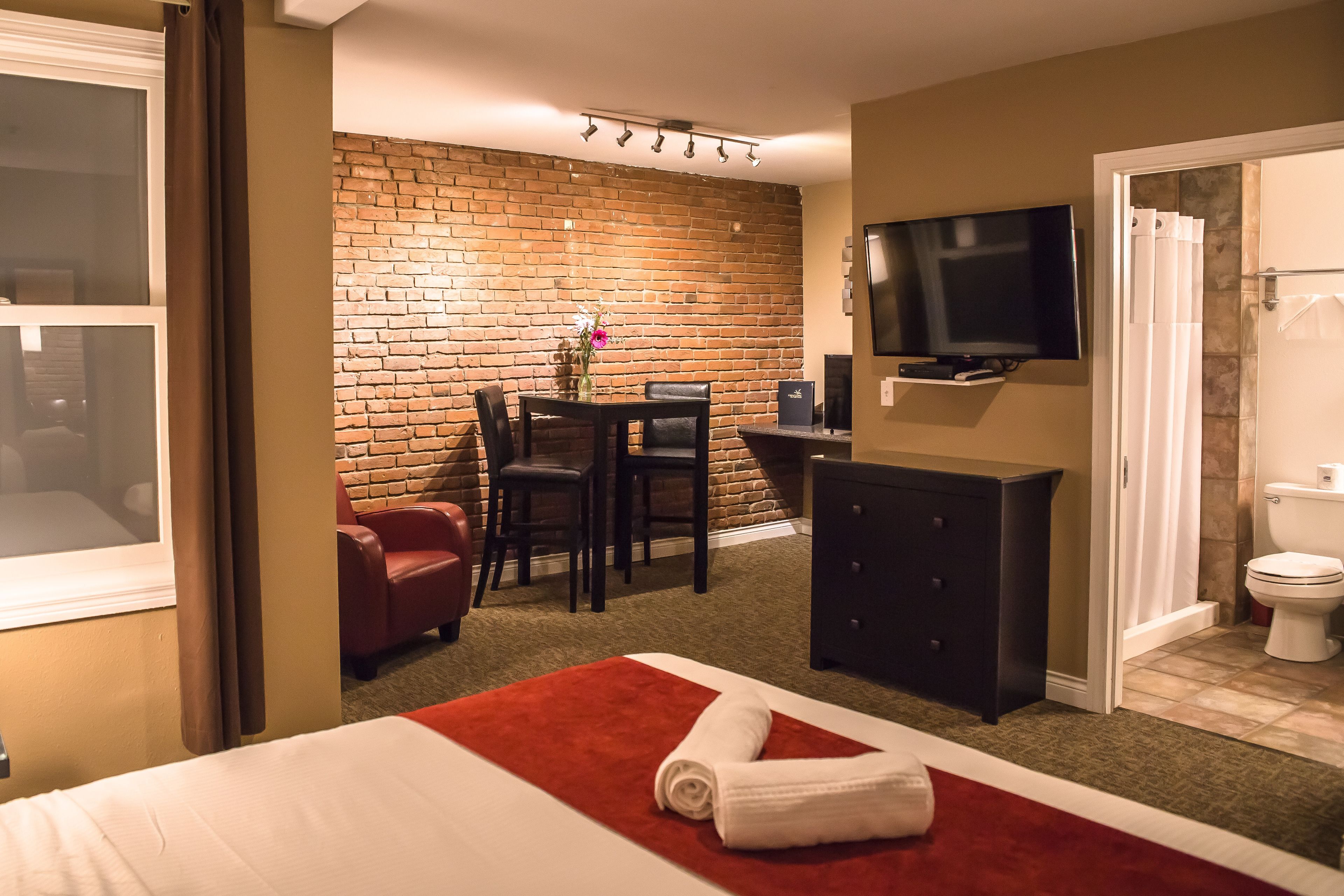 Una habitación de hotel con pared de ladrillo, televisión, mesa de comedor con sillas, alfombra roja, cama blanca y baño con inodoro y ducha.