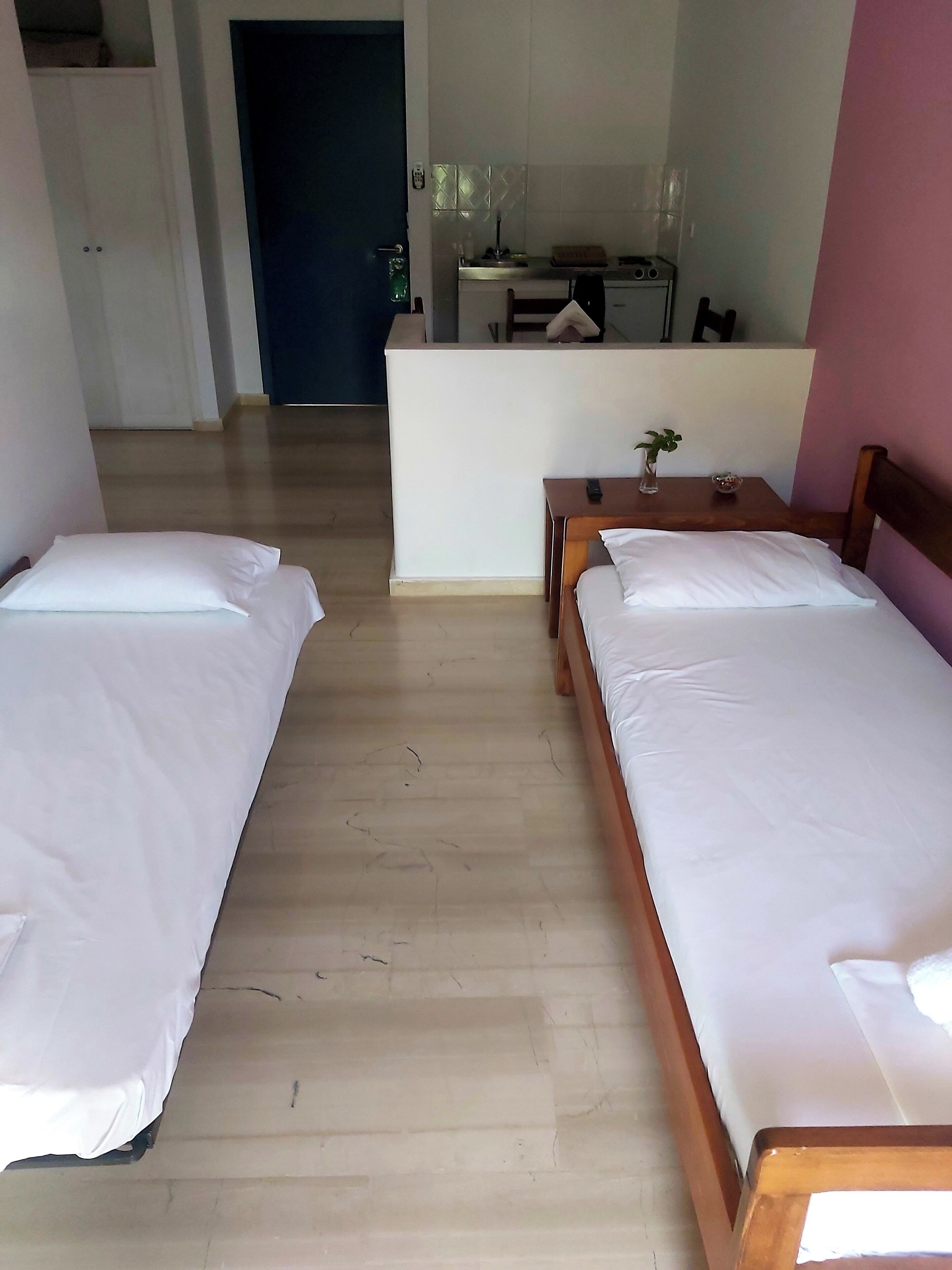 A hotel room with two single beds, a small table with a vase, and a kitchenette in the background.