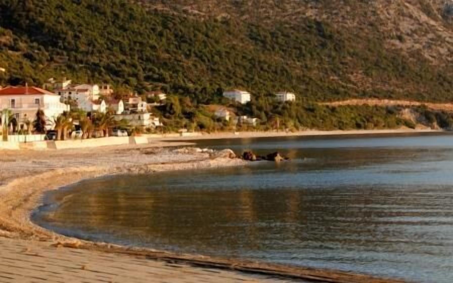 A coastal area with buildings near the water, a sandy beach, and a hilly background.