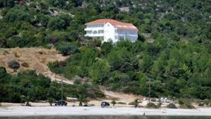 A white building with a red roof on a hillside, surrounded by dense greenery and a beach with a few cars. - Filoxenia Hotel & Apartments (Kefalonia)