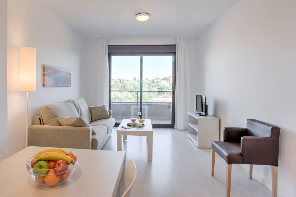 A modern living room with a sofa, a coffee table with fruits, a TV on a stand, and a view through a large window.