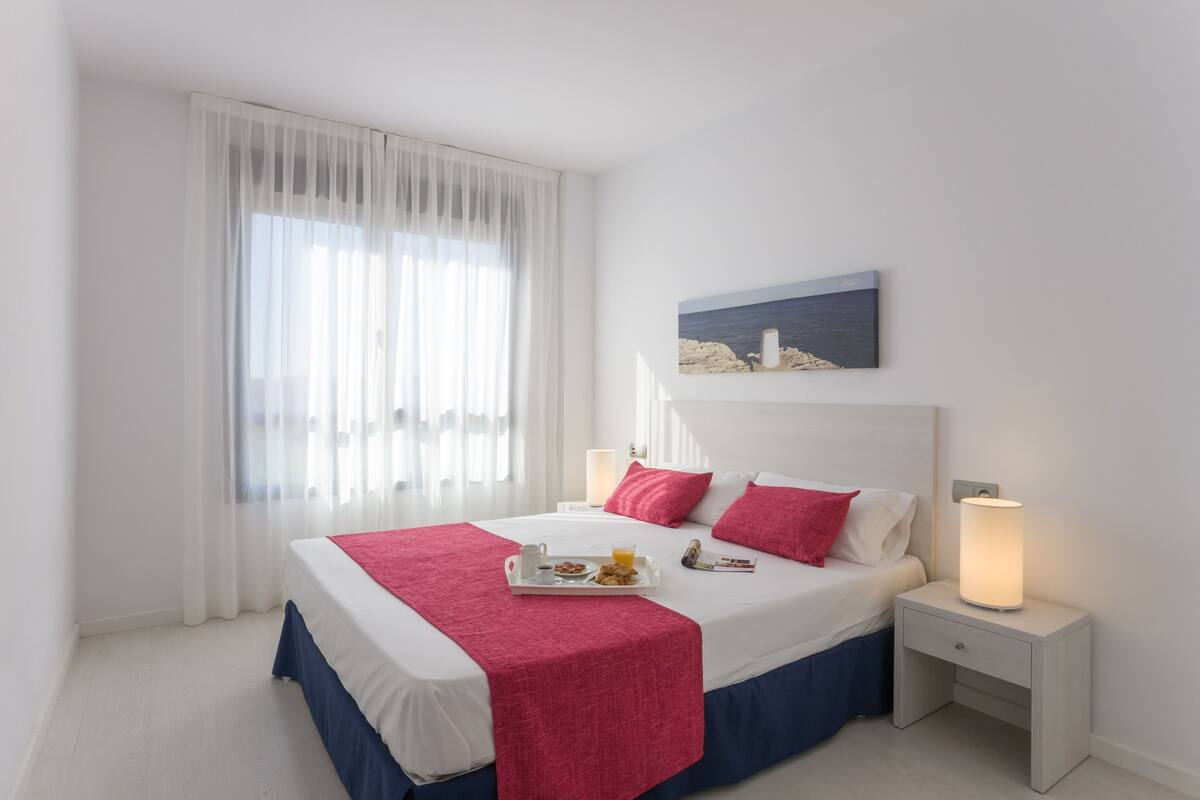 A neatly made bed with a red blanket, a tray with breakfast items, and a painting of a coastal scene on the wall.