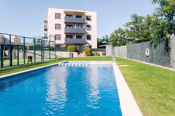 A swimming pool with a clear blue water, surrounded by a green fence, and a building with multiple balconies in the background.