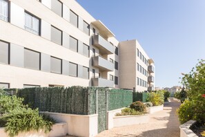 A modern multi-story residential building with balconies, a paved walkway, and landscaped areas with shrubs and flowers.
