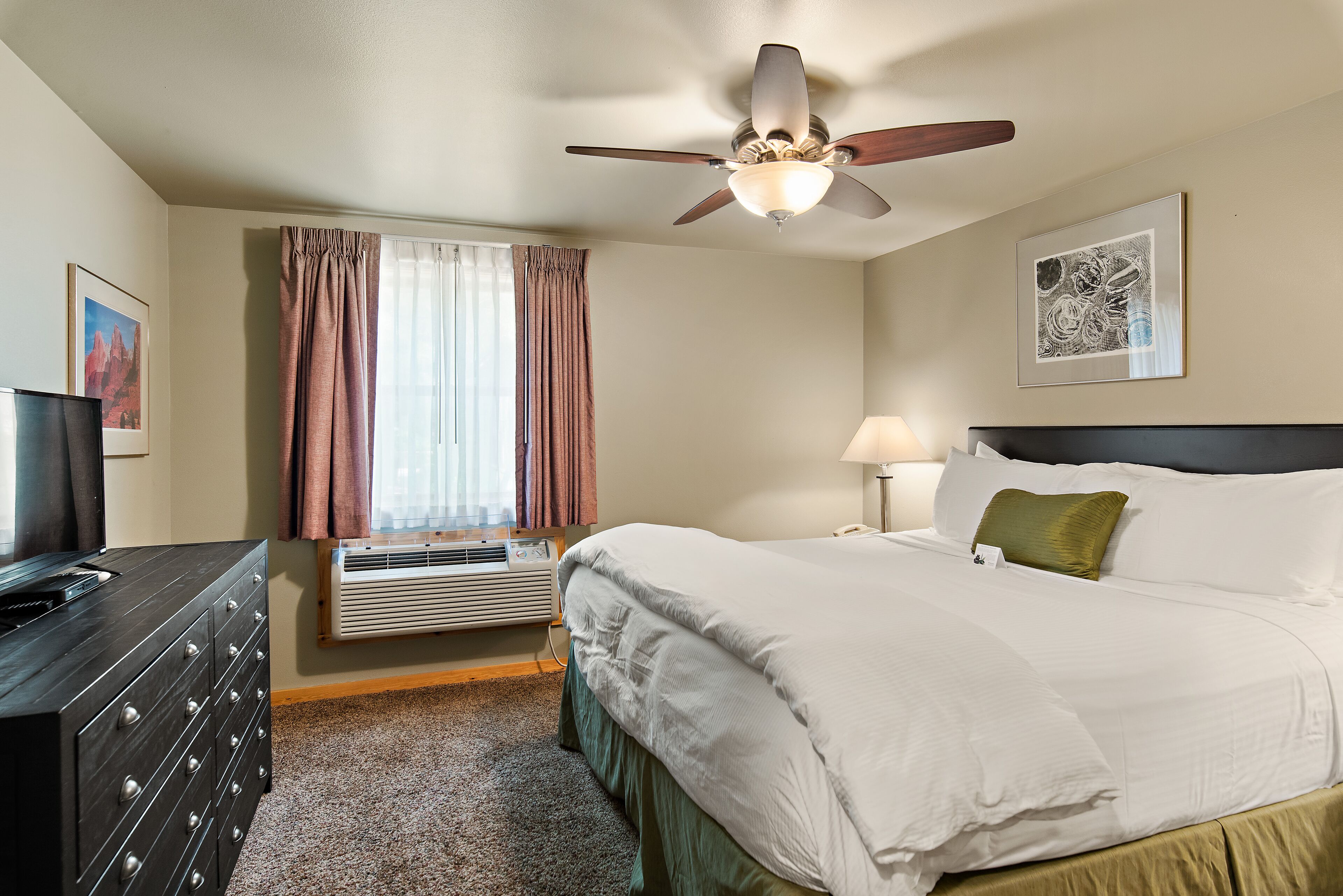 A hotel room with a bed, a ceiling fan, a window with curtains, a dresser, and a framed picture on the wall.