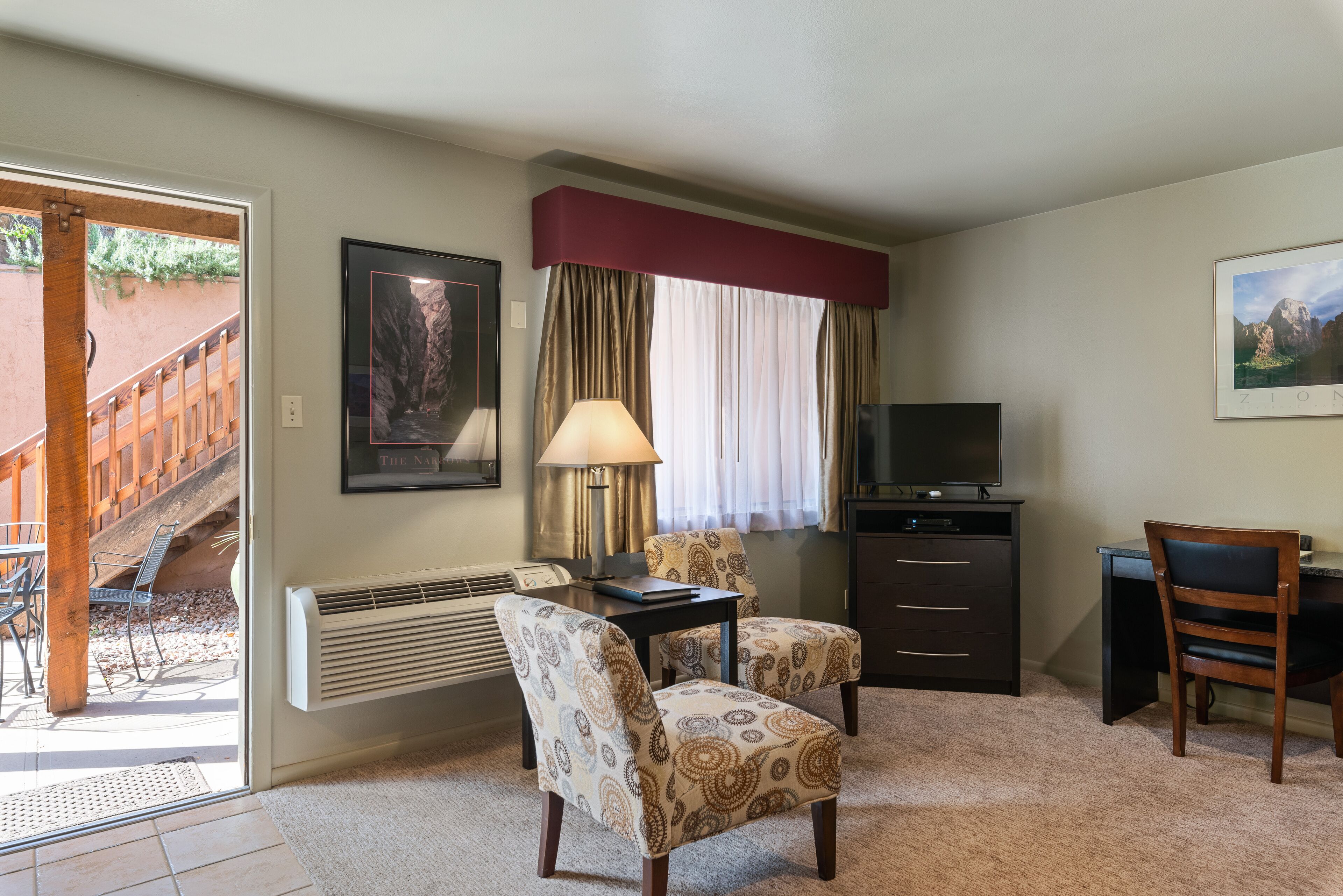 A room with a desk, two patterned chairs, a television, a heater, and a window with curtains.