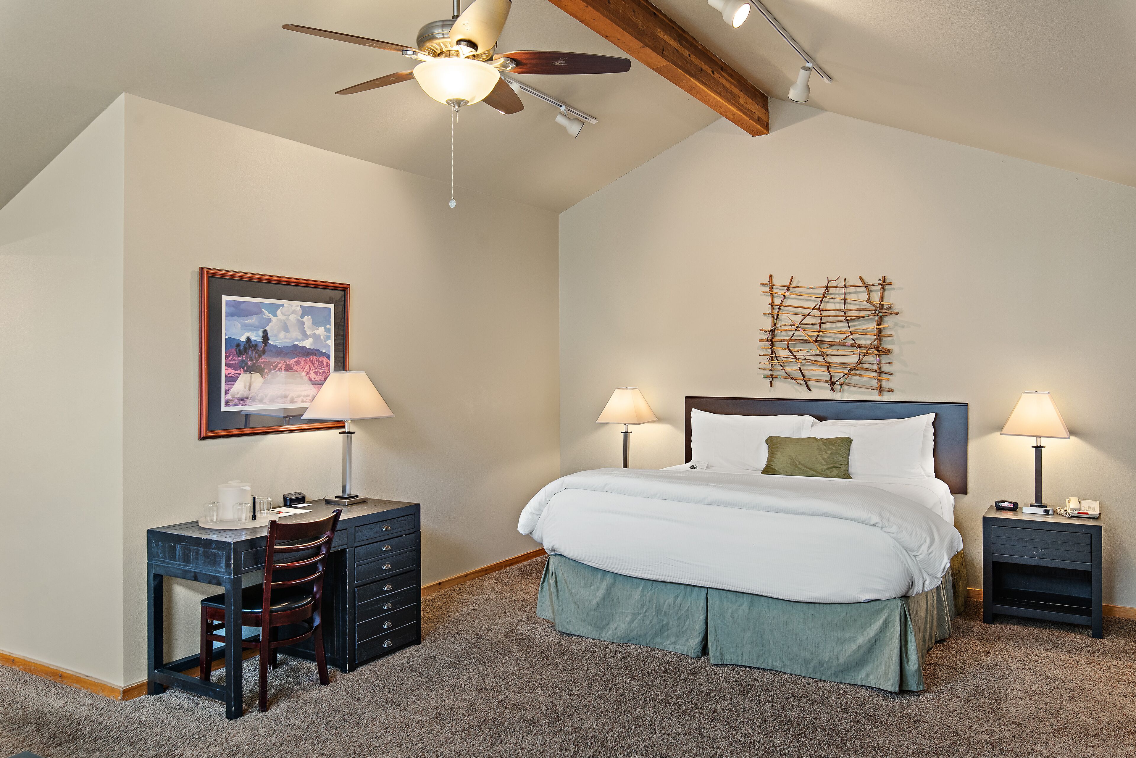 A bedroom with a bed, two bedside tables with lamps, a desk with a chair, a ceiling fan, and a framed picture on the wall.