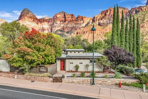 A hotel with a red door, surrounded by landscaped gardens and tall cypress trees, set against a backdrop of red rock formations.
