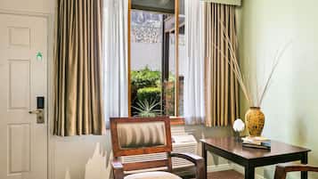 A hotel room with a wooden chair, a small table with a vase, and a view of a mountain through a window with curtains.