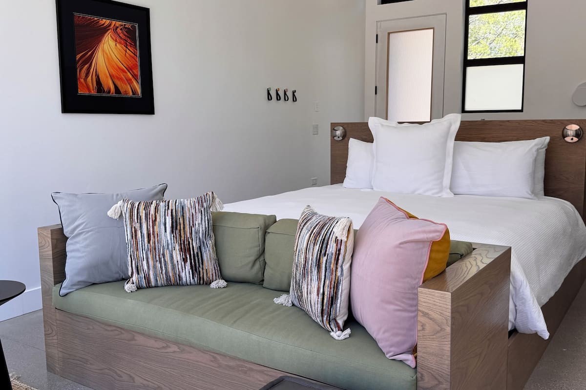 Een moderne slaapkamer met een bed, een groene bank met meerdere kussens, een klein poufje, een plafondventilator en een ingelijst schilderij aan de muur.