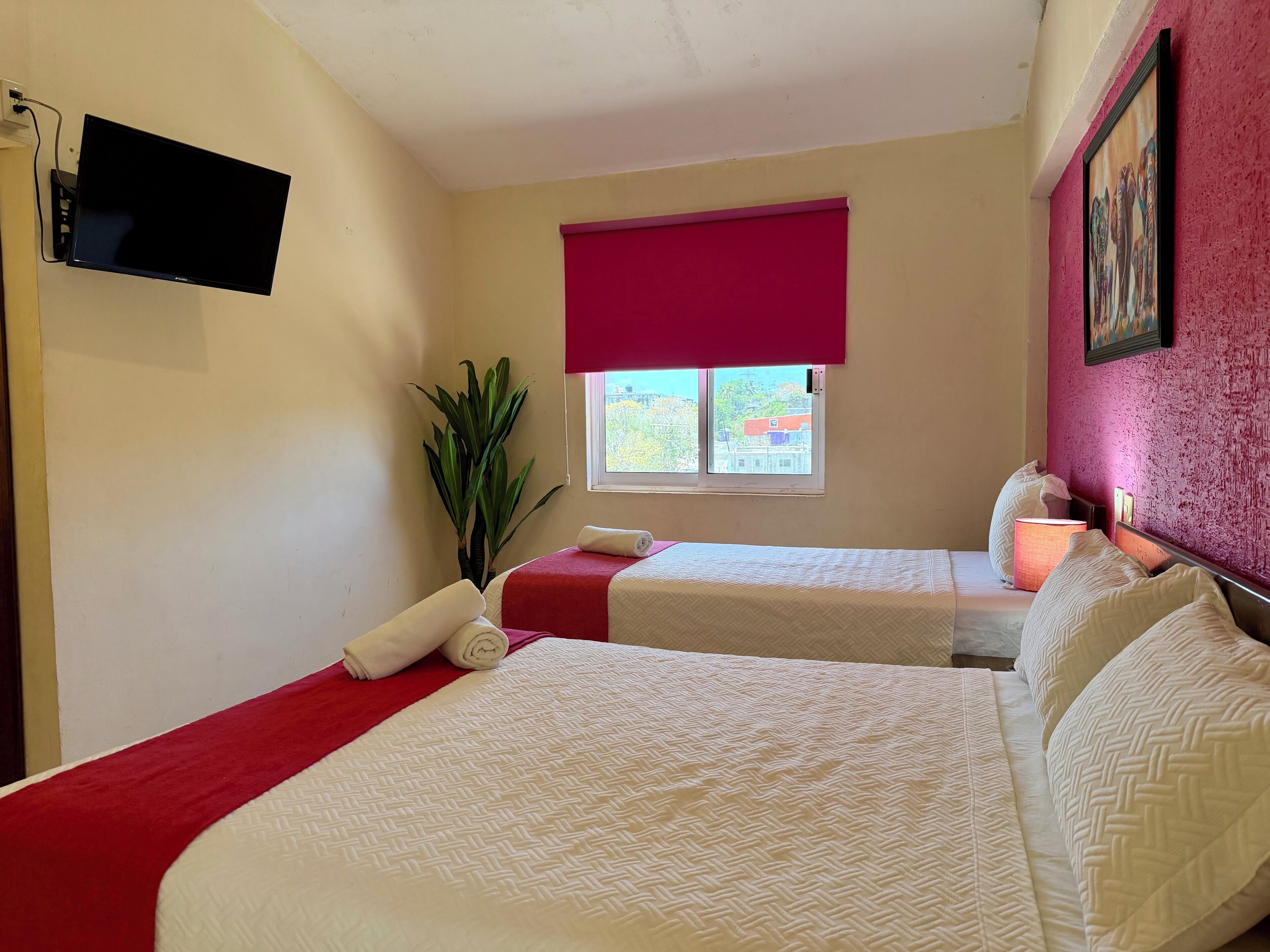 Habitación de hotel con dos camas, un televisor, una ventana con persiana roja, una planta y un cuadro en la pared.
