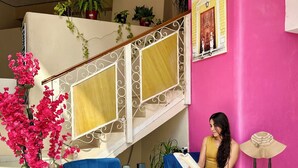 A woman sitting on a blue chair in a room with a pink wall, a staircase, and a potted plant with pink flowers. - Hotel Barranquilla (Campeche)