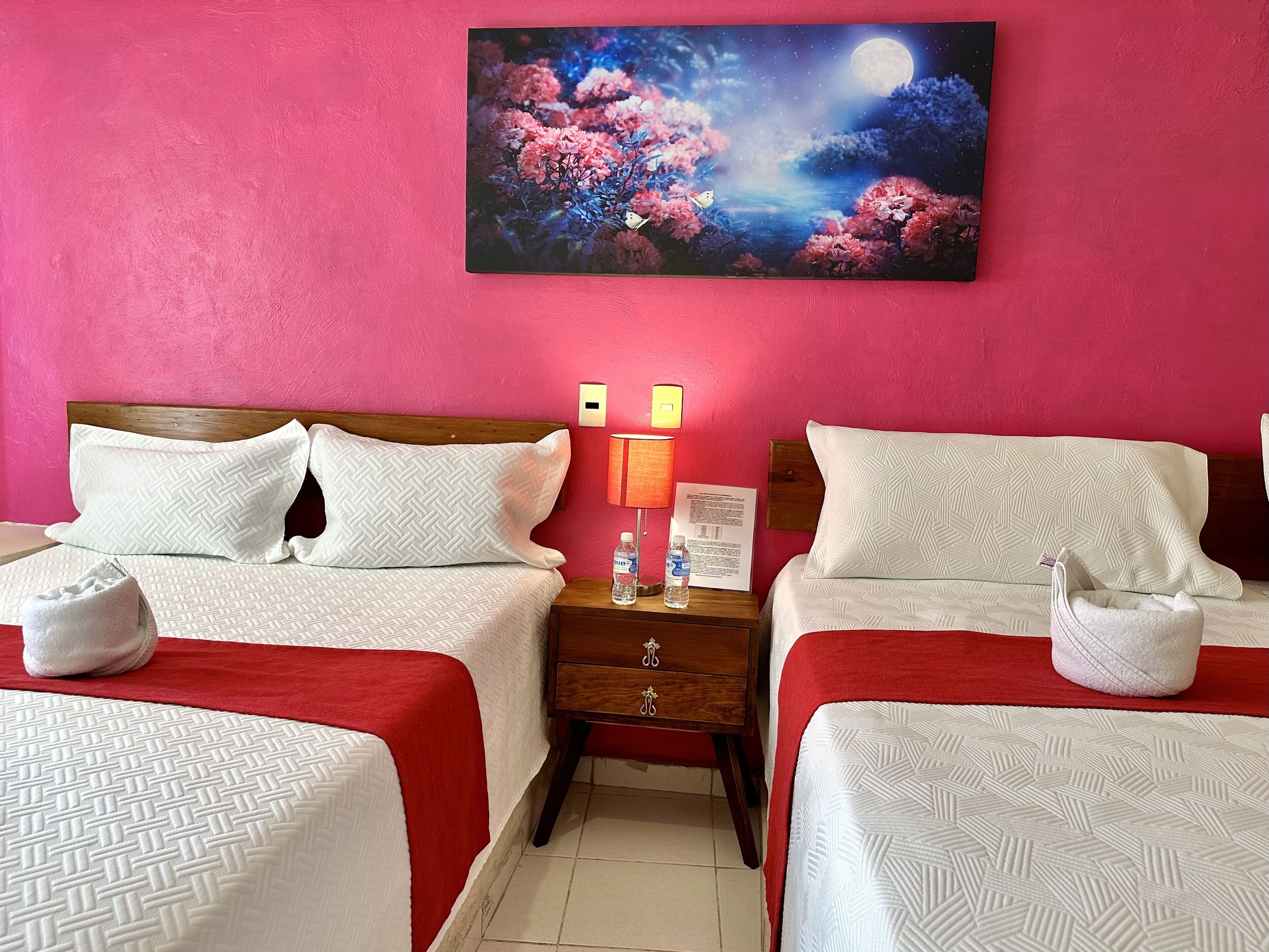 A hotel room with two beds, each with white bedding and a red blanket. There is a nightstand with a lamp, a water bottle, and a framed picture on the wall.