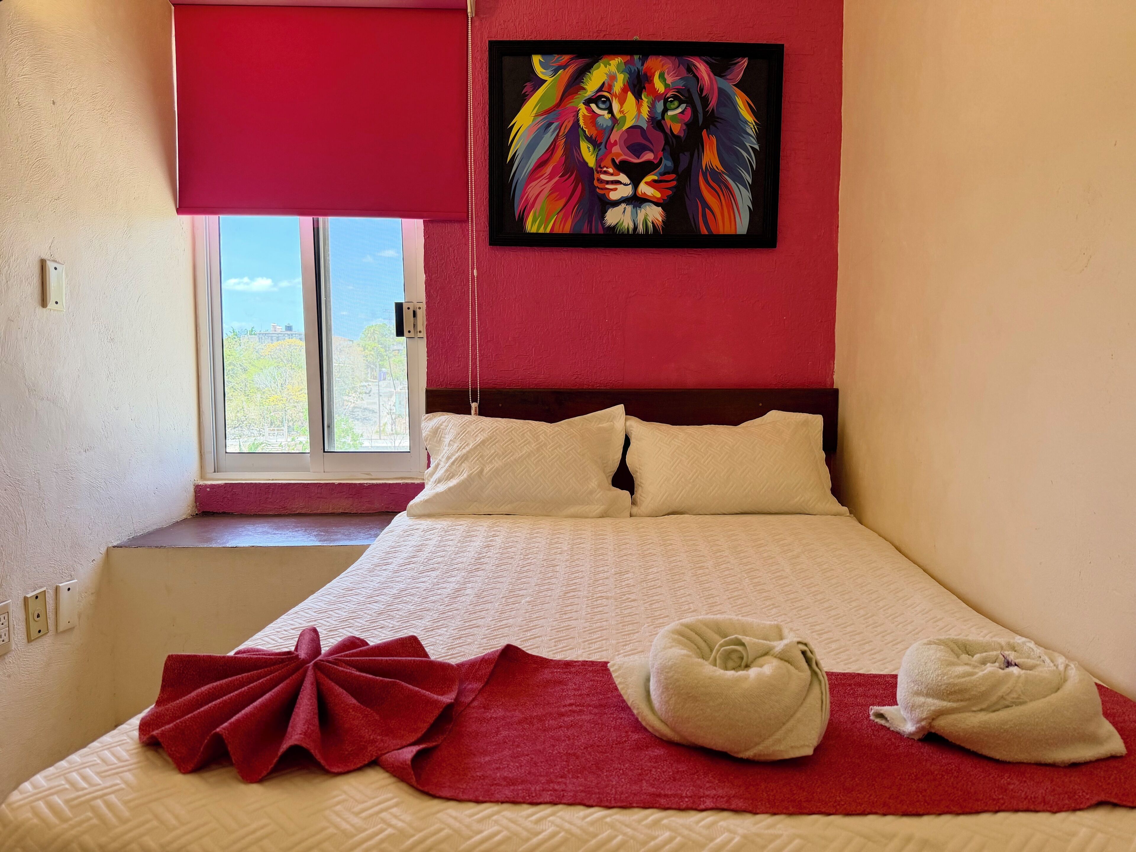 A bedroom with a bed, pillows, a red blanket, a towel, a window with a view, and a colorful lion painting on the wall.