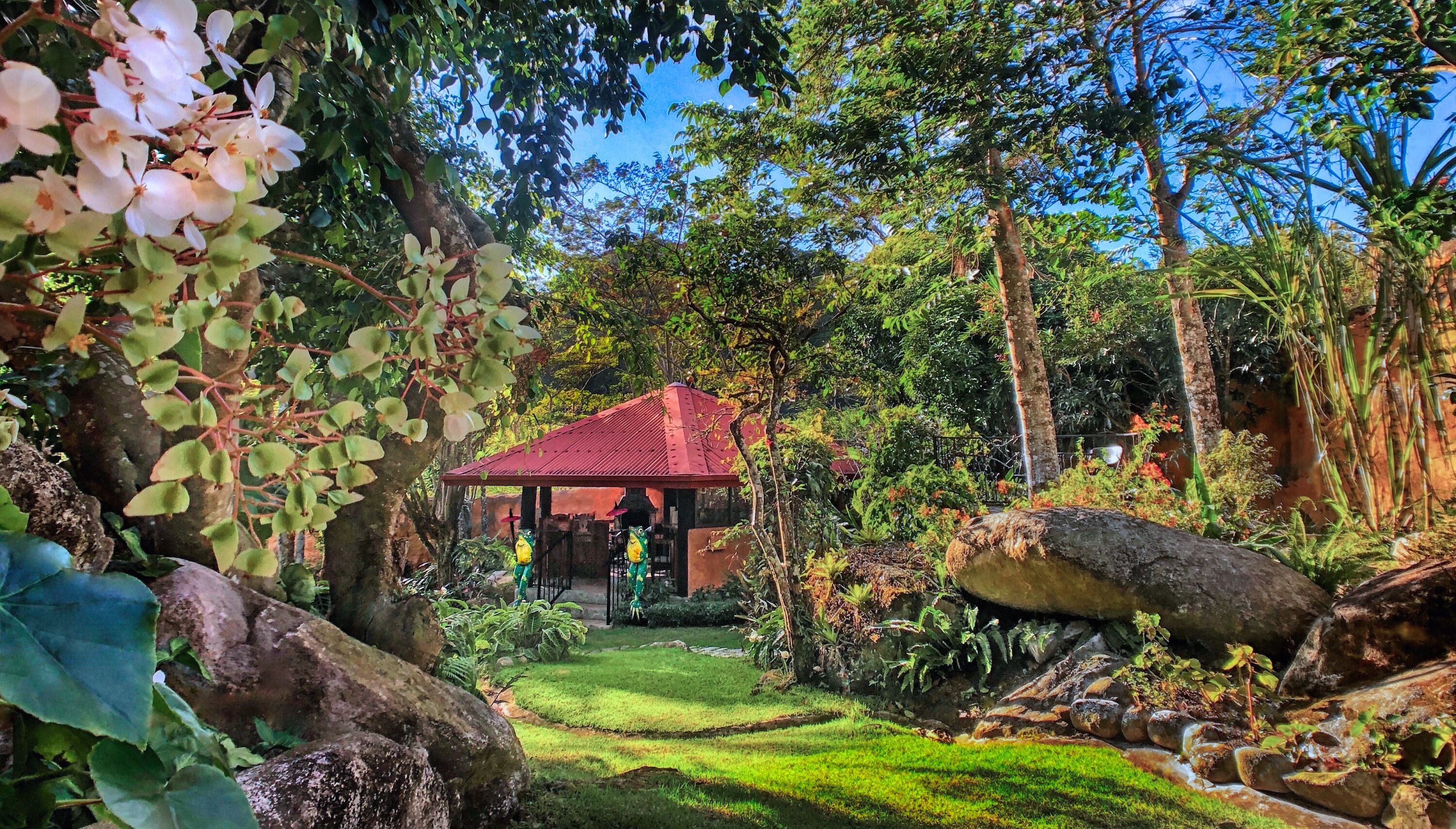 Un jardín con un pabellón, abundante vegetación y un pequeño arroyo.