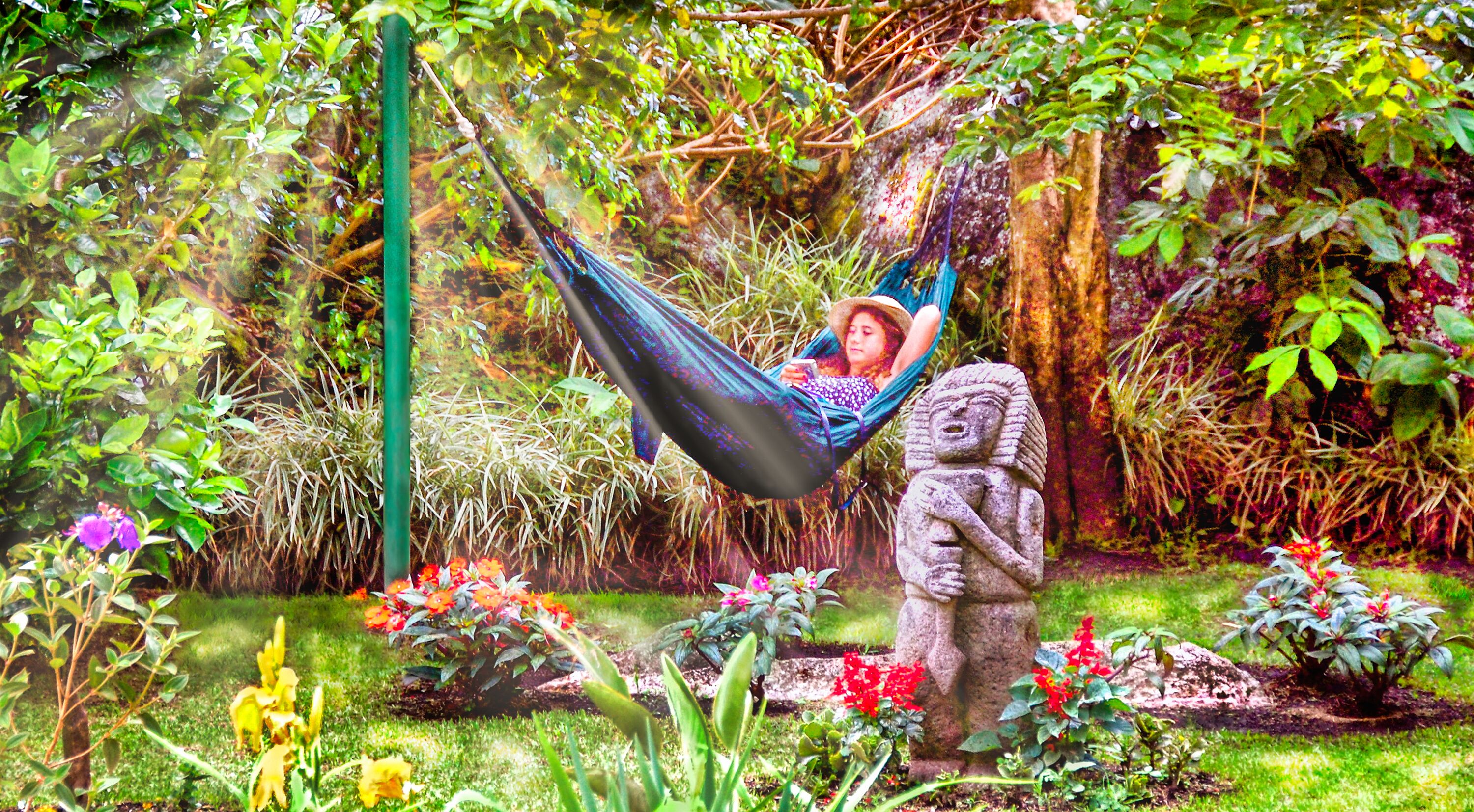 A hammock in a lush garden with a statue and various plants.