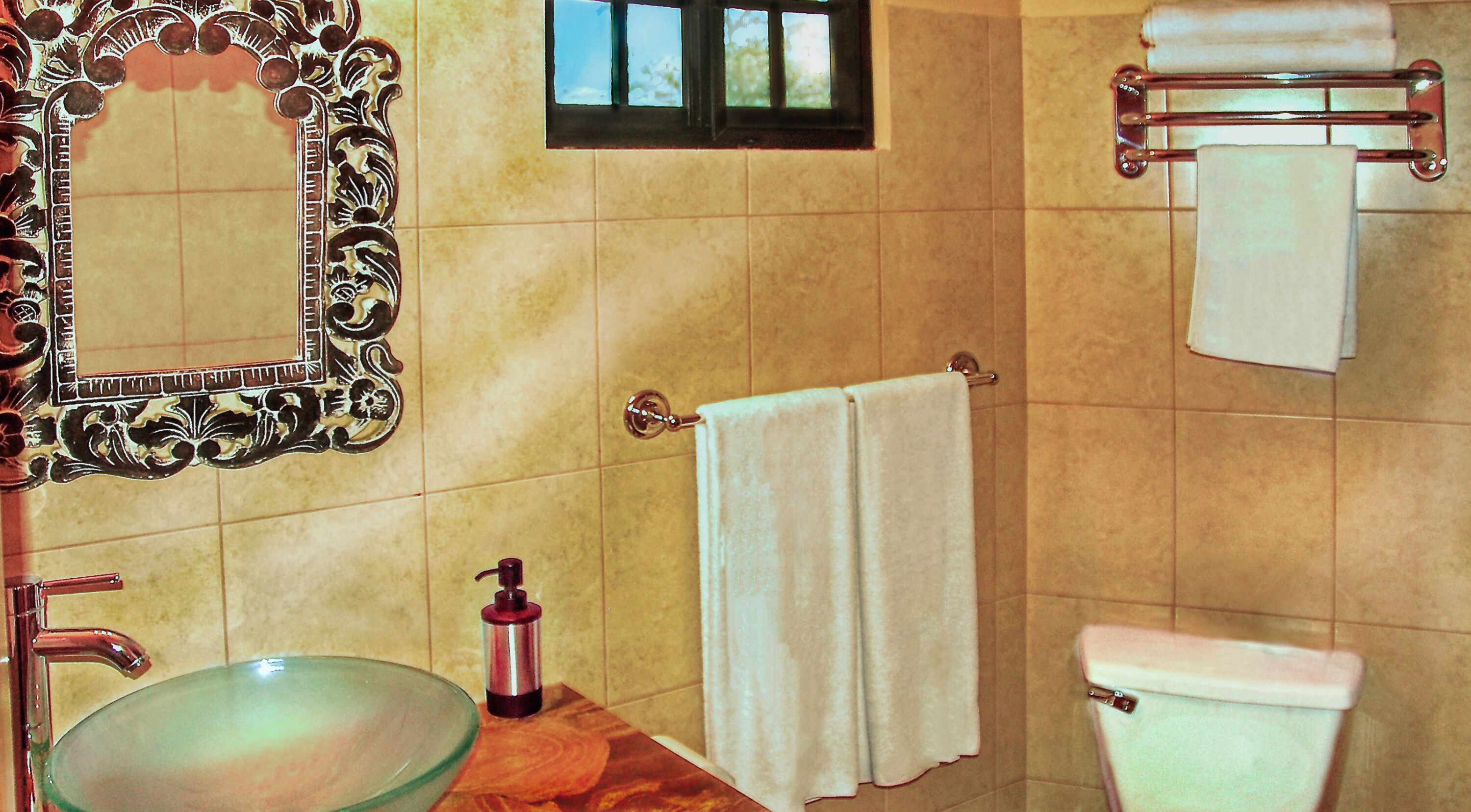 Een badkamer met een wastafel van glas, een decoratieve spiegel, een raam, een handdoekhouder en een toilet.