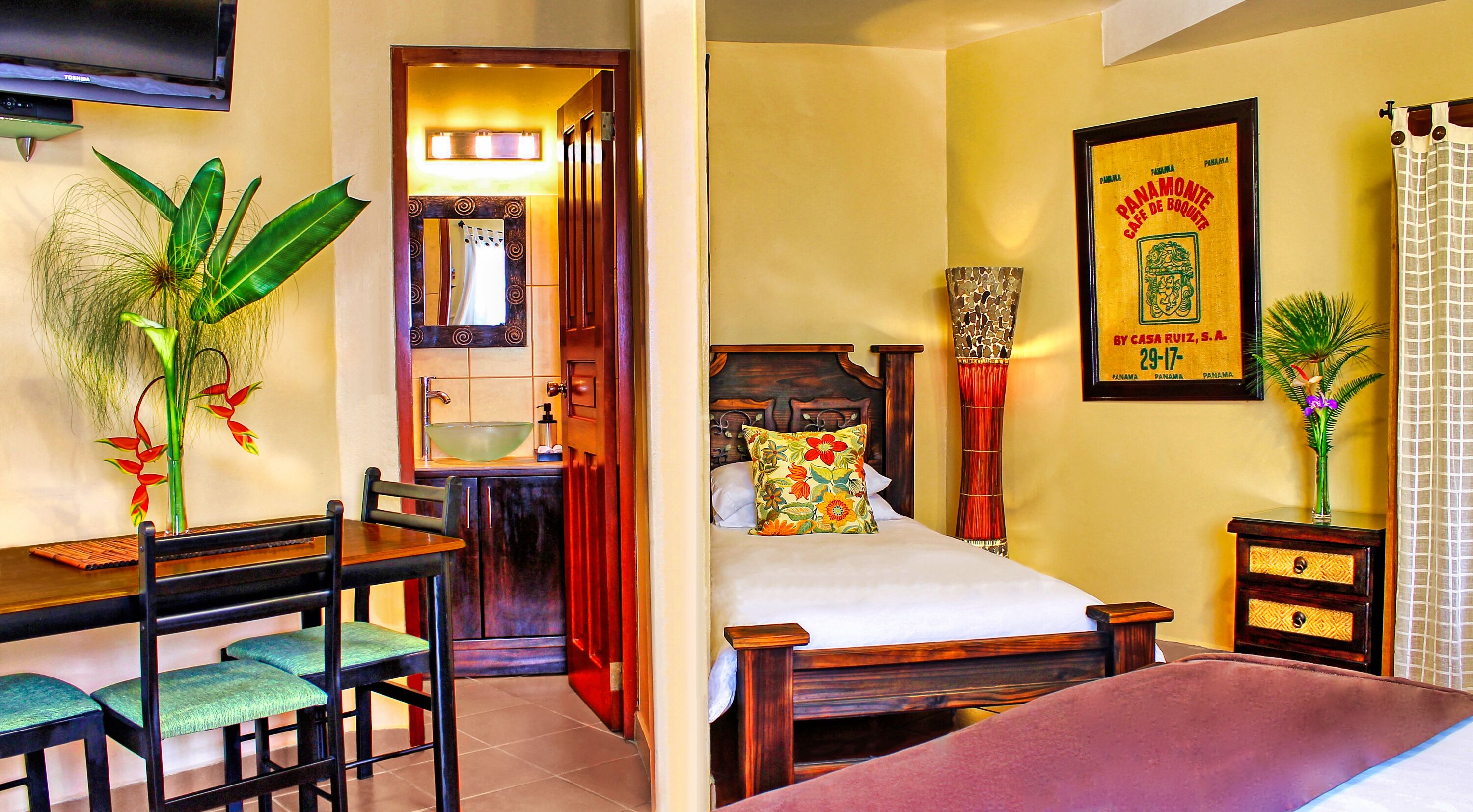 Une chambre d’hôtel comprenant un lit, un bureau avec des chaises, une salle de bain avec un lavabo et un miroir, et un tableau encadré accroché au mur.