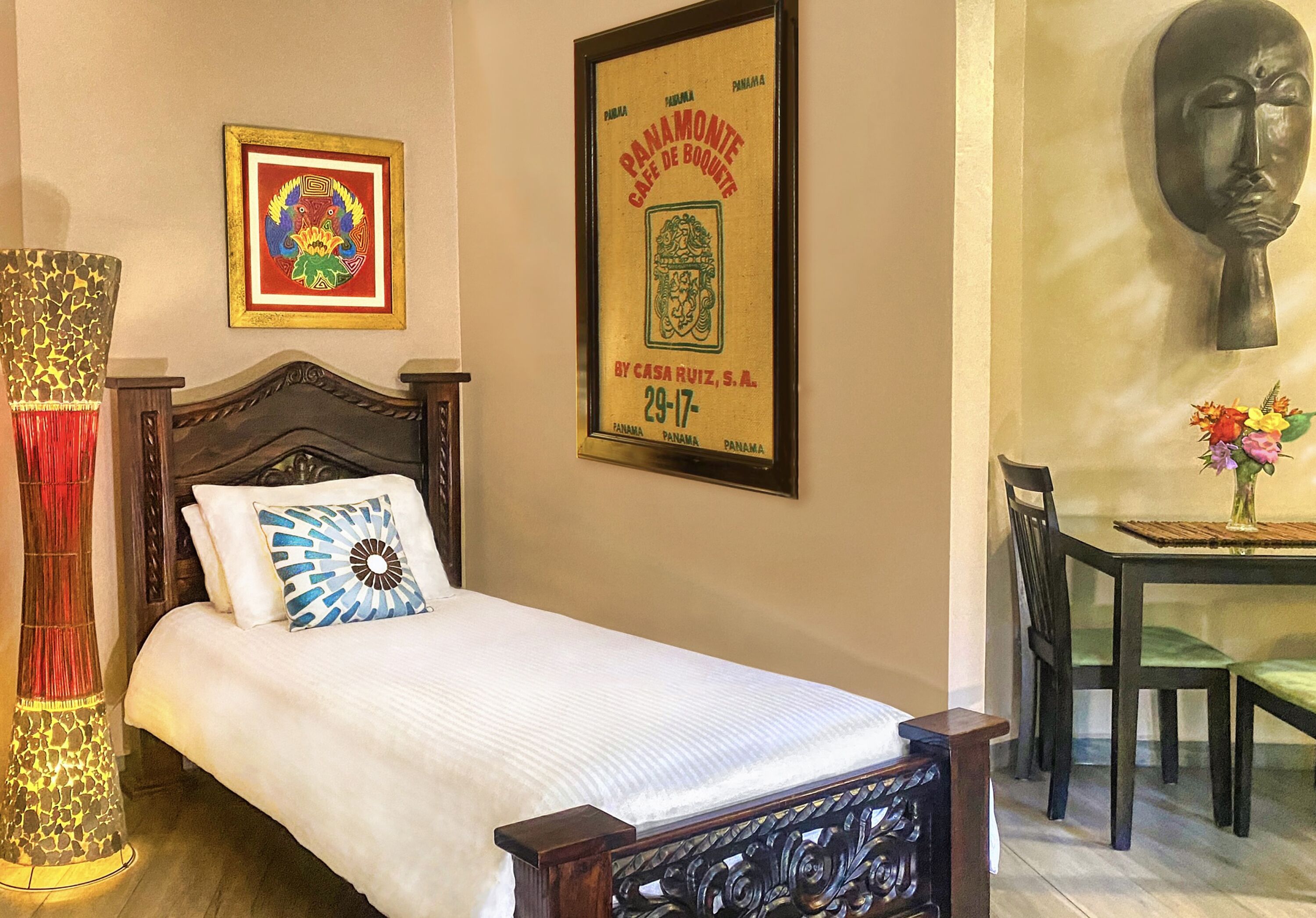 A neatly made bed with a white comforter and a blue and white pillow, a framed picture of a colorful emblem on the wall, and a framed menu for a Panamanian cafe.