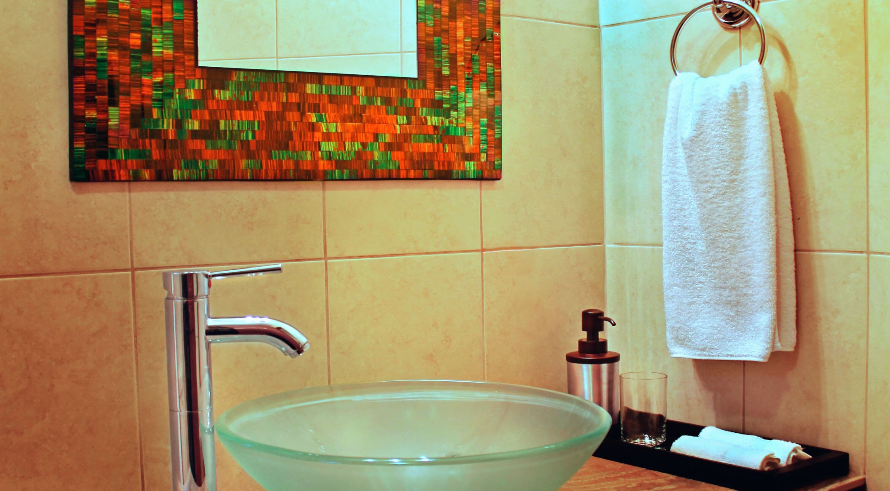 Een badkamer met een glazen wasbak, een chromen kraan, een zeepdispenser en een handdoek aan een rek.