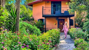 A two-story building with a balcony, surrounded by lush greenery and flowering plants.