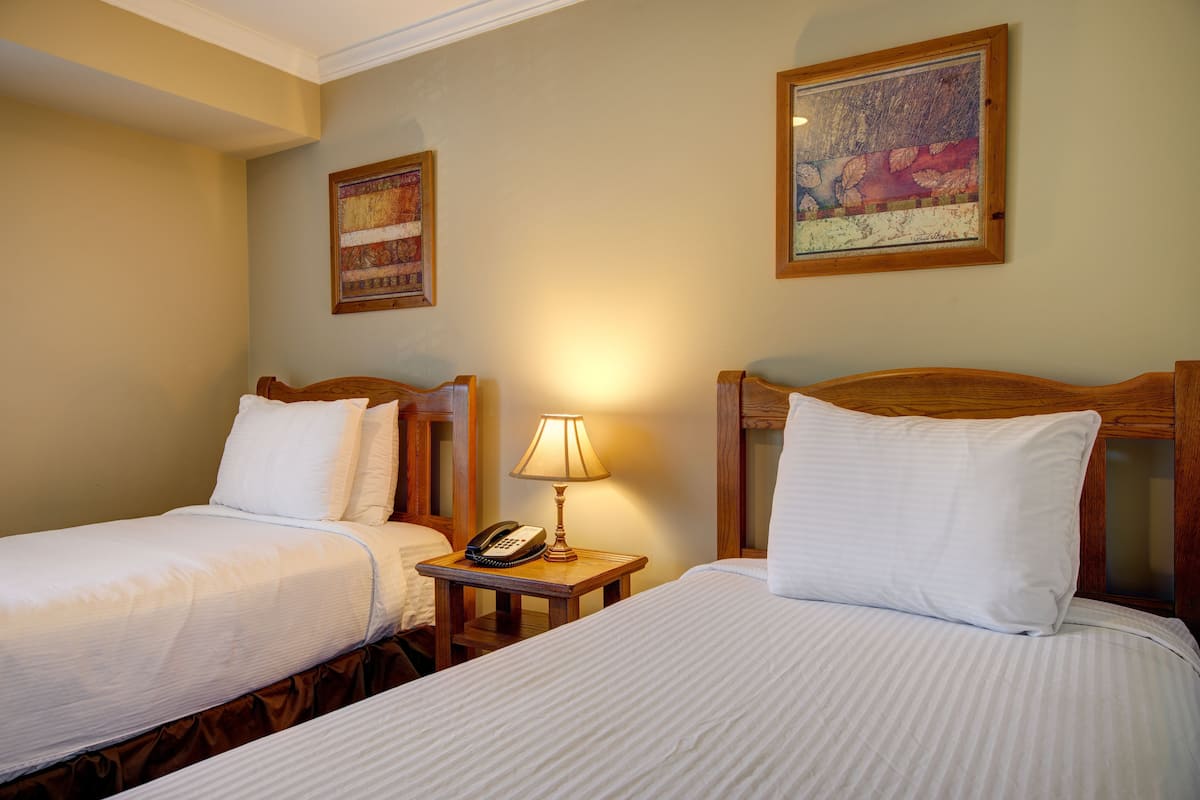 A hotel room with two single beds, a bedside table with a lamp, a phone, and two framed artworks on the wall.