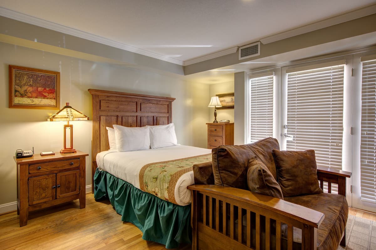 A bedroom with a wooden headboard, a green skirted bed, a wooden nightstand with a lamp, a wooden bench, and a window with blinds.