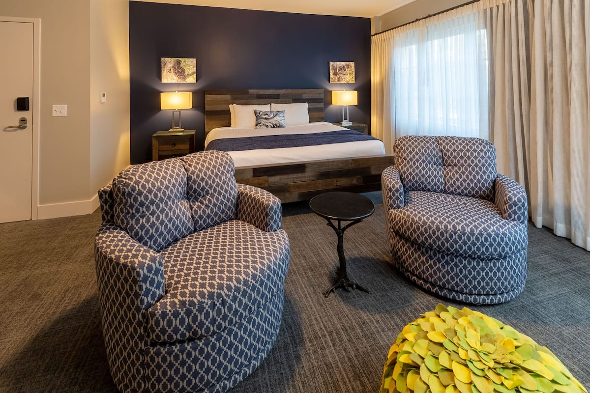 A hotel room with a bed, two patterned armchairs, a small table, and a decorative leaf arrangement.
