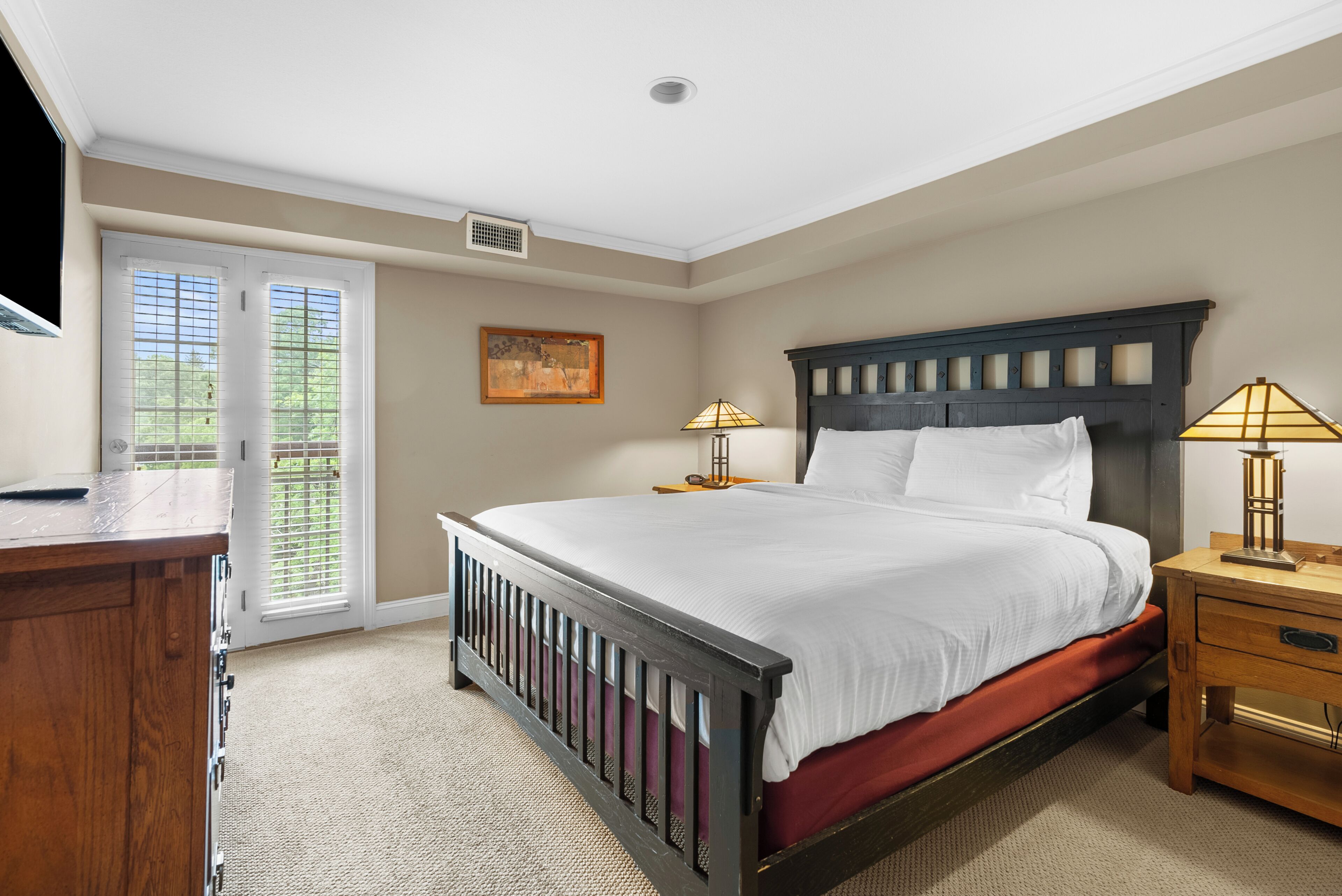 A bedroom with a bed, bedside tables, a desk, a chair, a window with blinds, and a painting on the wall.