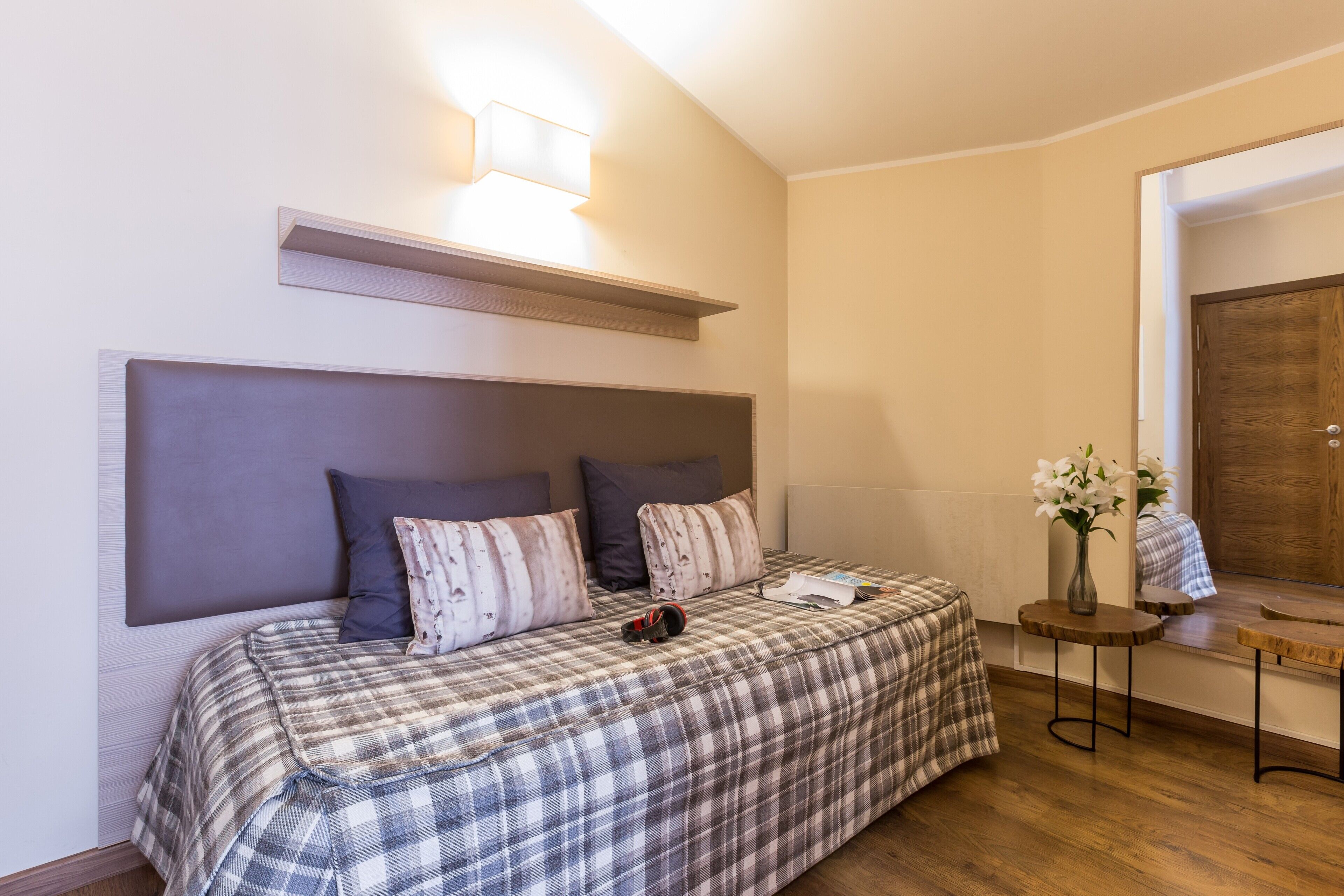 Un dormitorio con cama, almohadas, una mesita de noche con un florero y una puerta de madera.