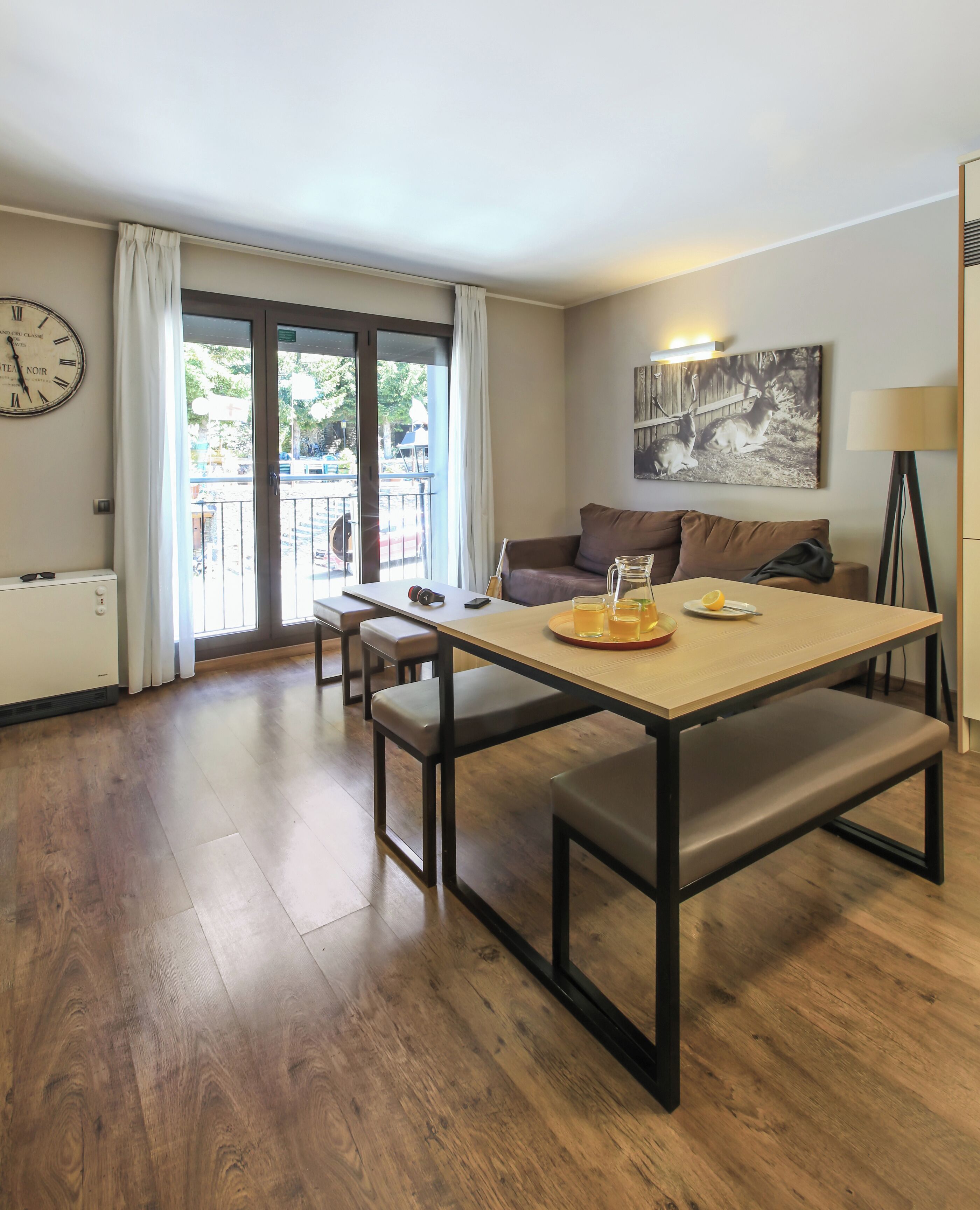 A modern living room with a wooden floor, a sofa, a dining table with benches, a clock, and a painting on the wall.
