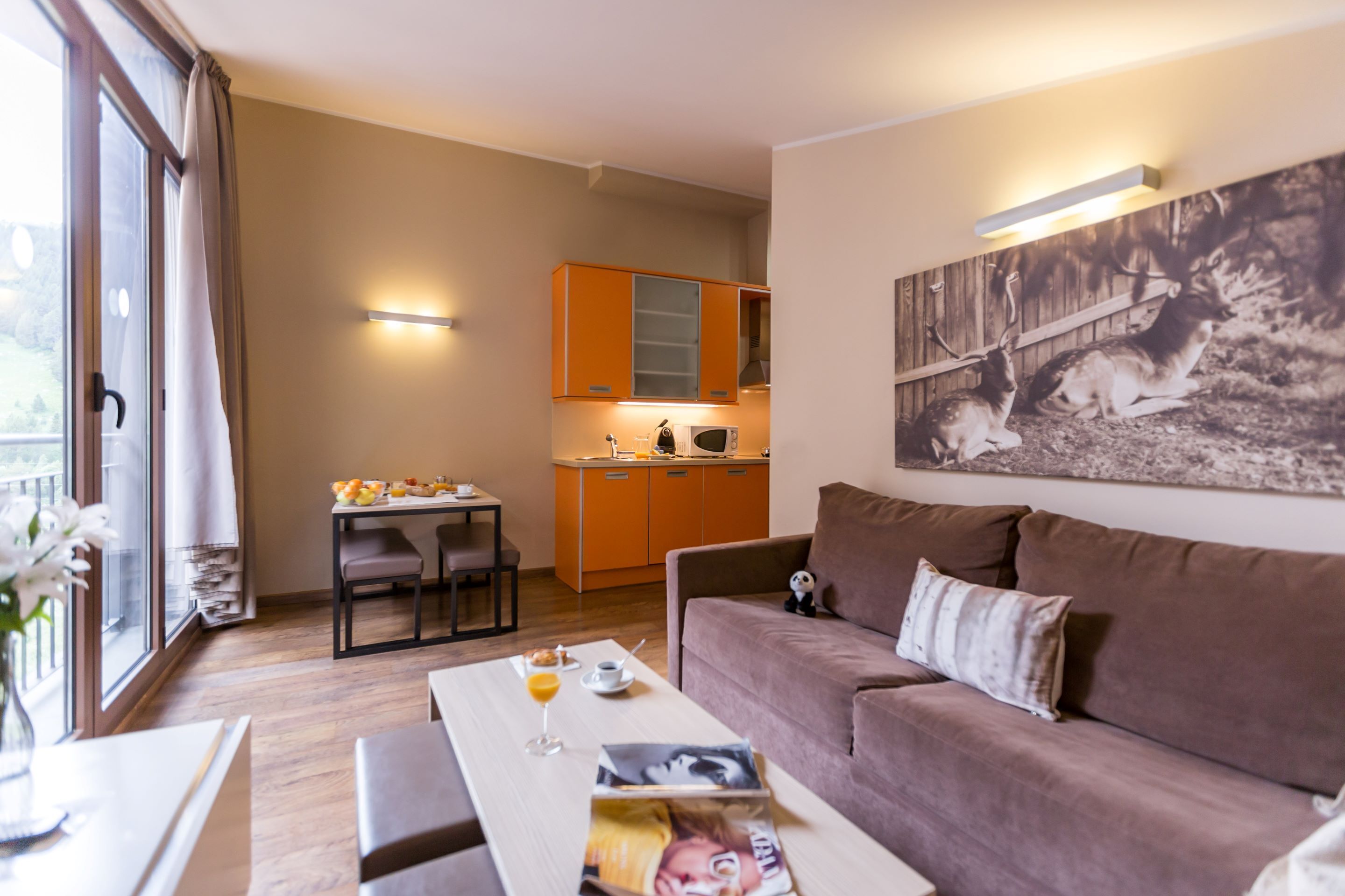 Ein modernes Wohnzimmer mit einem braunen Sofa, einem Esstisch mit Frühstücksutensilien, einer Küche im Hintergrund und einer großen Wanddekoration mit Darstellungen von Hirschen.