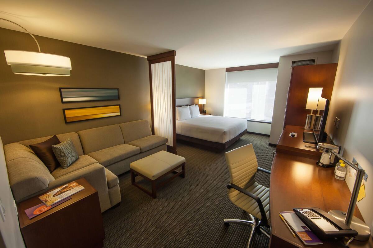 Une chambre d’hôtel avec un canapé, un lit, un bureau avec une lampe, une chaise et une fenêtre avec des rideaux.