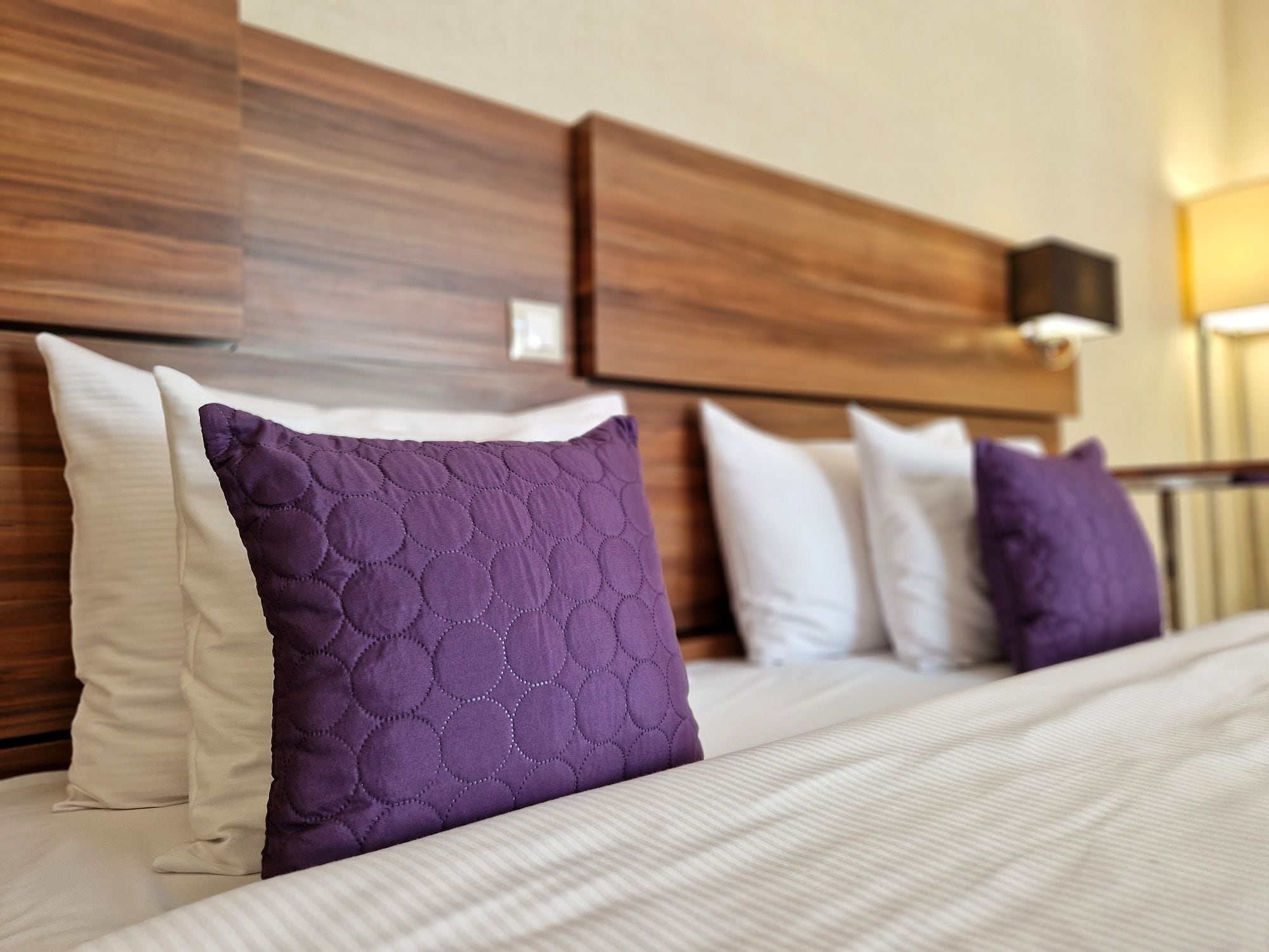 A hotel room with a wooden headboard, a bed with white and purple pillows, and a bedside table with a lamp.