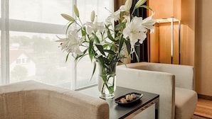 Een kamer met een groot raam, een vaas met witte bloemen, een kleine tafel met eten erop, en een lamp.
