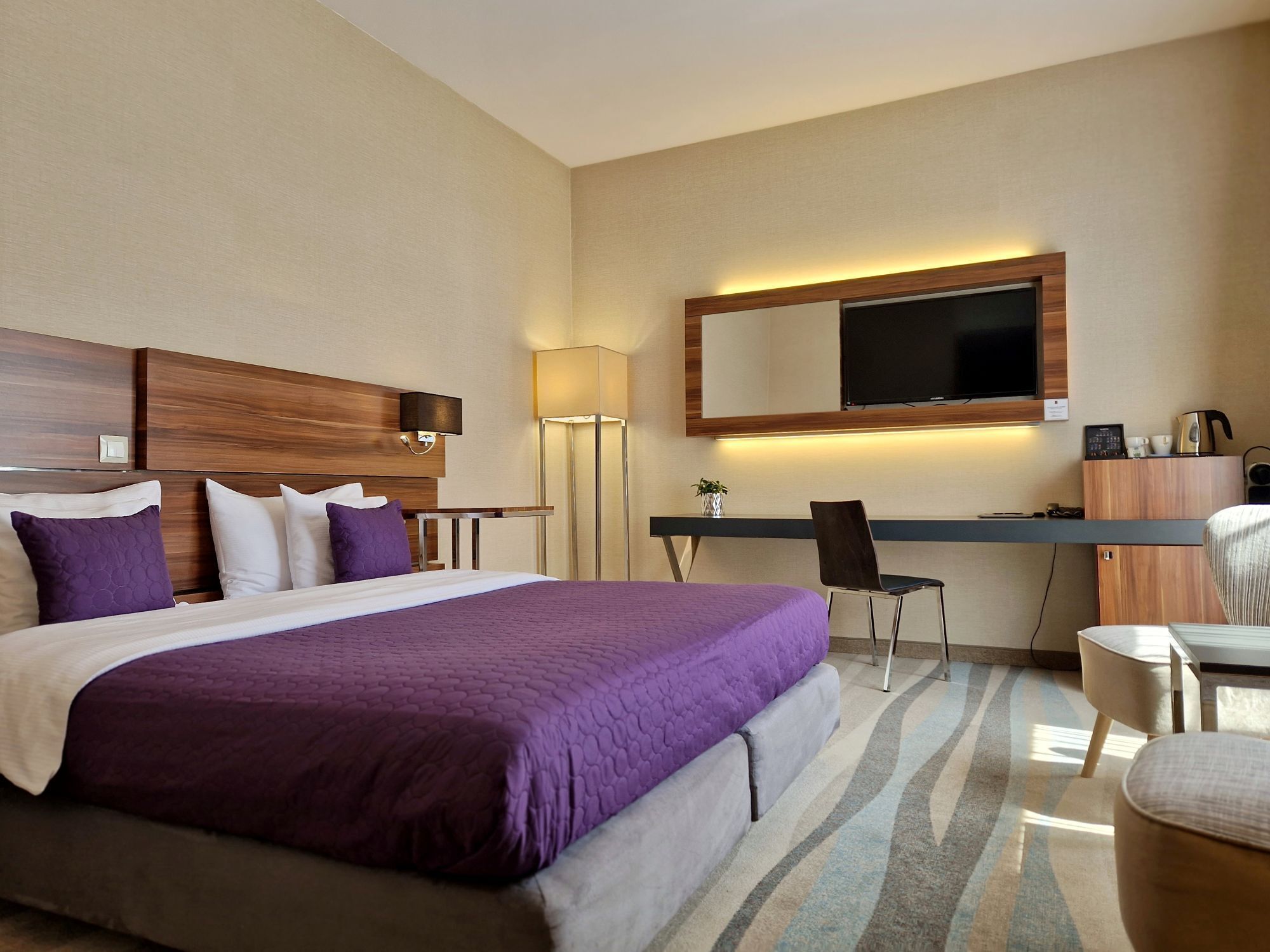 A hotel room with a large bed, a desk with a chair, a TV, and a lamp.