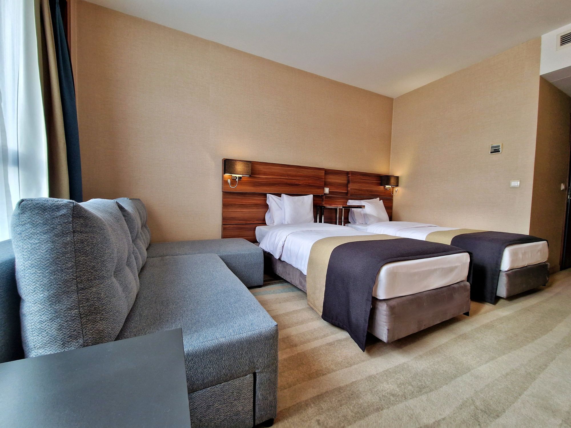 Une chambre d’hôtel avec un canapé, deux lits, une tête de lit en bois et une lampe fixée au mur.